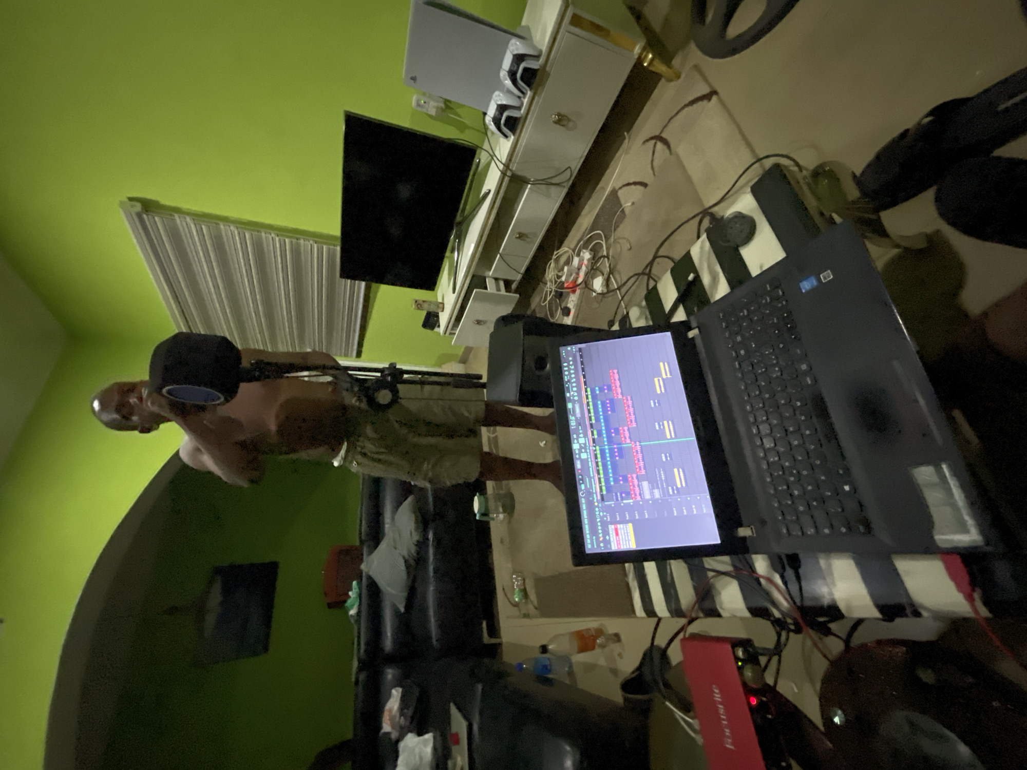 The image depicts a person in a room that appears to be a makeshift music studio. The individual is light skinned, bald, and shirtless, wearing shorts and appears to be standing in front of a microphone. A laptop displaying a music production software interface sits nearby, next to a red audio interface. The room is characterized by green walls and contains a TV, a console with a PlayStation 5, a couch, and various pieces of equipment and cables strewn around. The location is not definitively indicated as being in Abuja, Nigeria, but the scene suggests a casual, home-based studio setup. The image does not provide information regarding the individuals' actions or interactions with others.