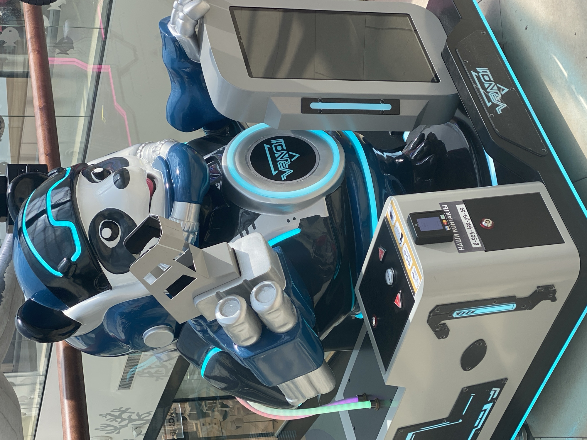 A close-up, eye-level, medium shot captures a futuristic, life-sized robot panda amusement ride, possibly a VR experience, situated indoors. The panda, dressed in a blue and silver robotic suit with glowing neon blue accents and trim, sits behind a control panel. Its right arm is extended, holding a metallic device, while its left arm rests on a gray console. The console features a screen and various buttons, some triangular and red, others round and white, labeled with Cyrillic text. A prominent sign on the console reads "НАШИ КОНТАКТЫ" (Our Contacts) followed by a phone number.
The base of the ride is black, adorned with a neon blue light strip and the "VRN01" logo. To the right of the panda is another gray console with a vertical blue light. The ride appears to be located in a public space, possibly a shopping mall, with glass railings and blurred background elements suggesting an indoor environment. The lighting is bright, likely from overhead sources. No individuals are visible interacting with the ride. The overall impression is of a clean, modern, and technologically advanced entertainment attraction. The location is specified as Novokuznetsk, Russia.
