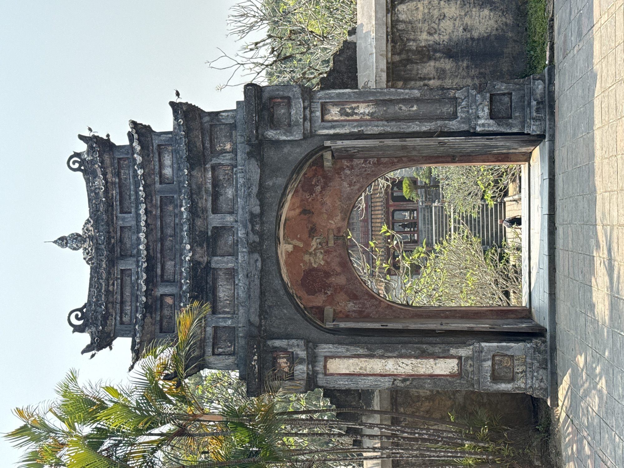 The image depicts an ornate stone archway, with birds perched atop its roof, in Kim Long Ward, Vietnam. The archway leads to a courtyard with a building and stairs. The archway's structure is aged and textured with decay. Palm trees and other greenery are visible near the archway. A paved walkway is on the right of the frame.