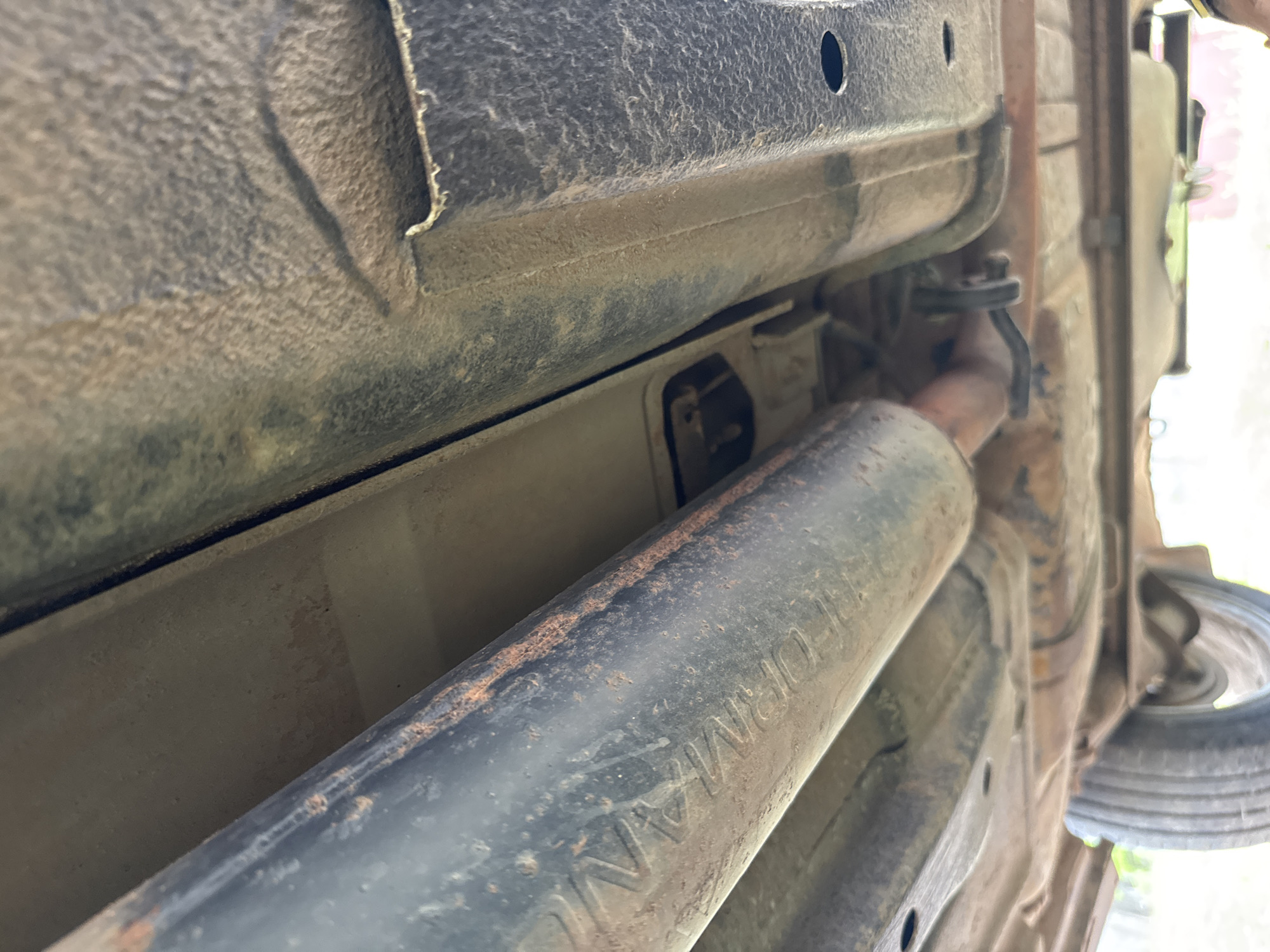 The image displays a close-up view of the underside of a vehicle, captured from below. The primary subject is a large, black, cylindrical exhaust component, likely a muffler or resonator, positioned diagonally across the foreground. This component shows noticeable signs of rust and dirt accumulation. Embossed or stenciled on its surface, the word "PERFORMANCE" is clearly visible.
To the left and above the exhaust component, various structural elements of the vehicle's undercarriage are visible, including metal beams and panels, some with a rough, textured finish, possibly from an undercoating. Further back and to the right, an exhaust pipe is connected to the frame via a hanger or bracket. On the far right, a spare tire is partially visible, suggesting the rear of the vehicle.
The lighting is bright, indicating it is daytime. No specific activity is depicted, but the vantage point suggests the vehicle is either elevated on a lift or positioned over an inspection pit, implying an inspection, maintenance, or repair context. The overall condition of the undercarriage and exhaust components shows wear, dirt, and some rust, consistent with a vehicle that has seen use. There are no other visible texts or specific location cues within the image itself, apart from the provided context of Бородулино, Russia.