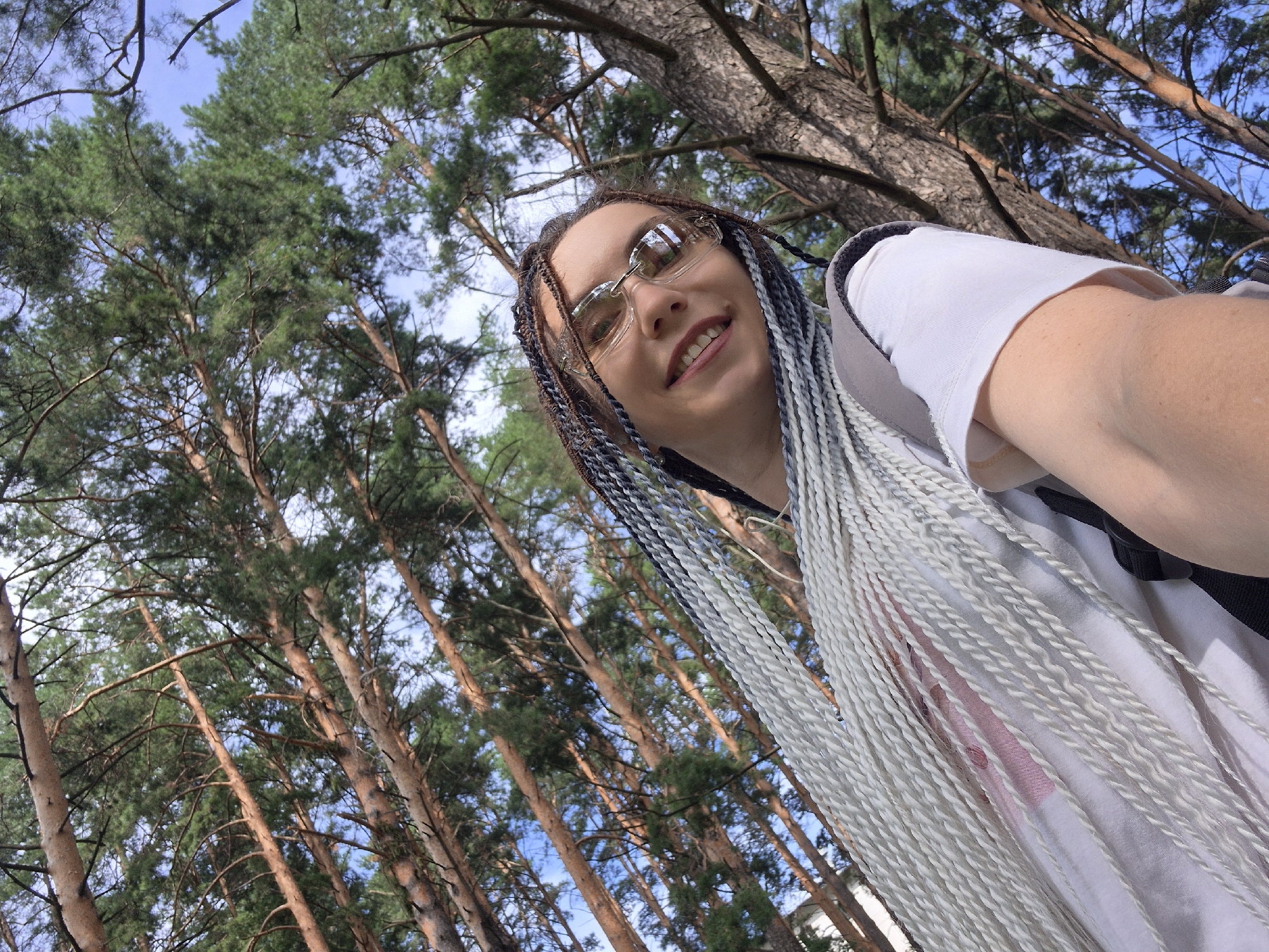 The image is a low-angle, selfie-style shot taken outdoors. A young woman with long, braided hair, predominantly white with streaks of gray and brown, is the primary subject. She is wearing glasses and a white t-shirt, with a hint of pink fabric visible underneath. She is smiling and looking directly at the camera.
The background is dominated by a canopy of tall pine trees with thin trunks and branches, characteristic of the coniferous forests found in Chaikovsky, Russia. The sky is visible through the branches, appearing blue with some scattered clouds. The lighting suggests it is daytime, with sunlight filtering through the trees, creating dappled shadows. The overall impression is of a person enjoying nature in a forested area.