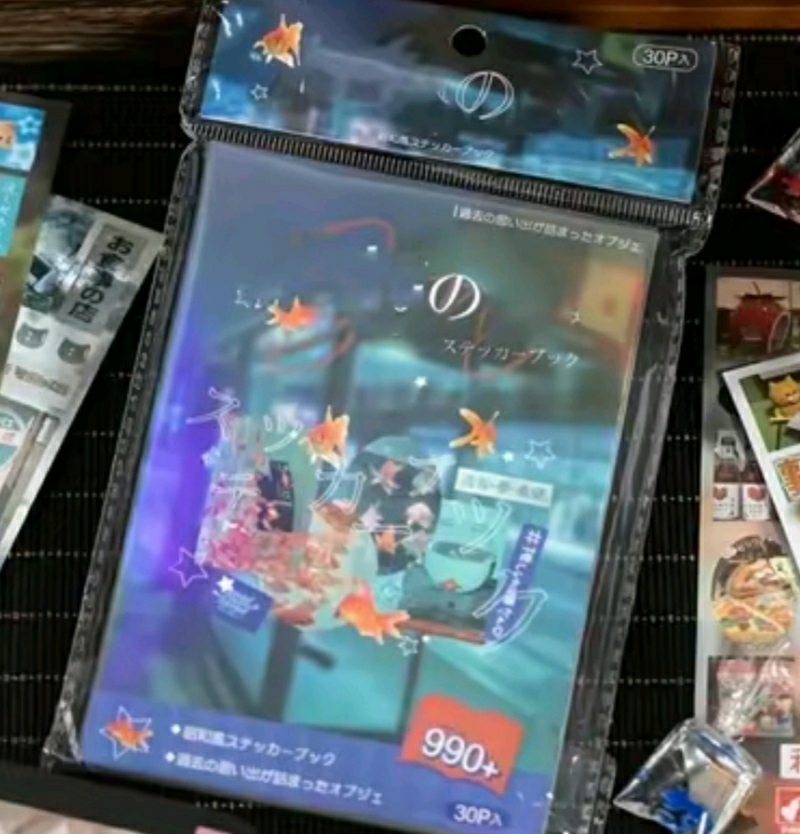 The image shows a sticker book in its packaging. The book features a blue color scheme with illustrations of goldfish and text in Japanese. The packaging includes the words "30PA" and "990+". There are other sticker packs around the main one. The image appears to be taken indoors on a dark surface with a woven pattern. There is no information about the location being in Minsk, Belarus.