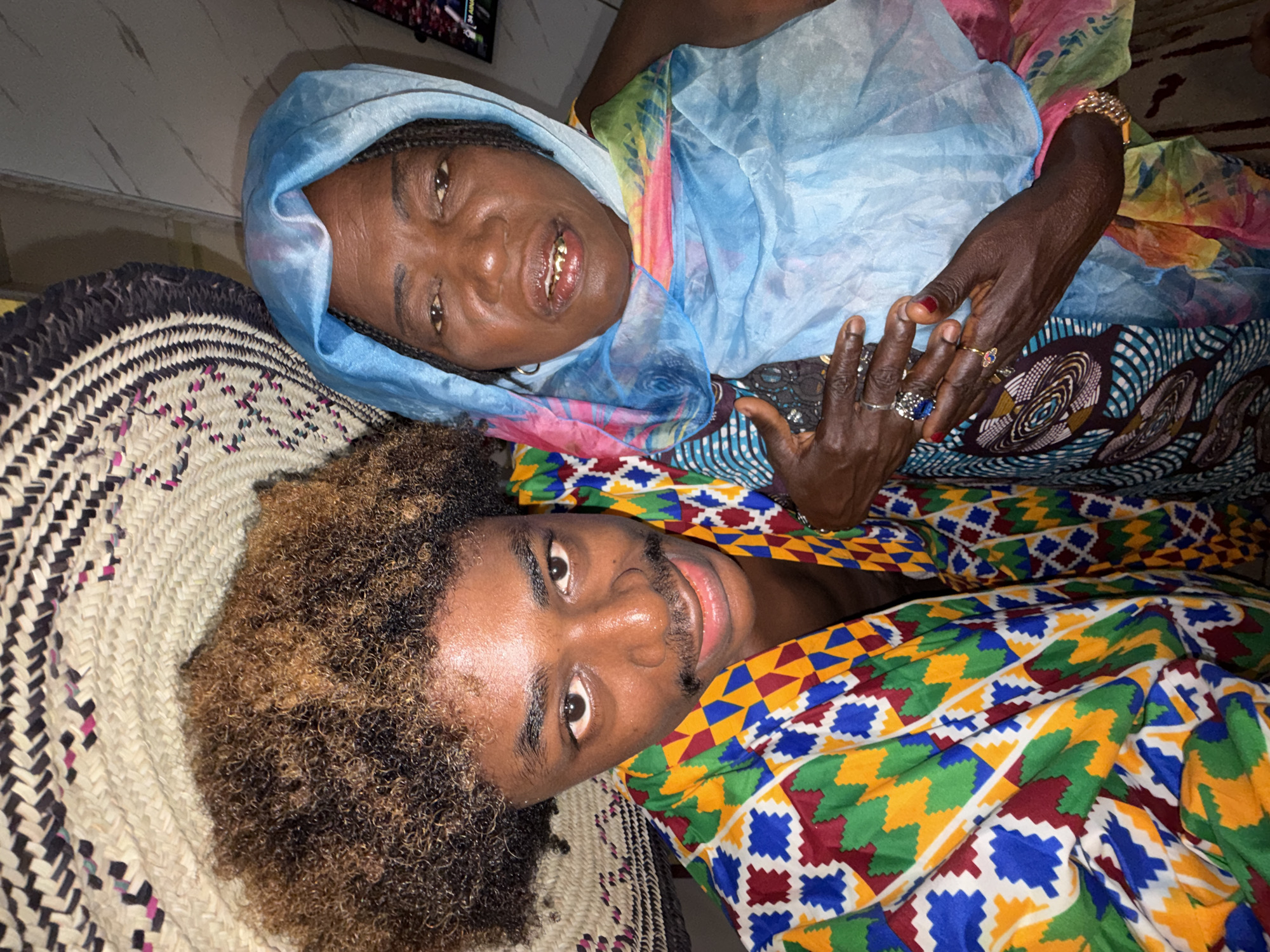 The image captures an overhead view of two individuals, an older woman and a younger man, lying side-by-side and smiling at the camera. The scene takes place indoors, likely in Igboefon, Nigeria, as indicated by the location context and cultural elements.
The woman, positioned on the left from the viewer's perspective, has dark skin and braided hair. She wears a light blue, translucent head covering that flows over a vibrant, multi-colored patterned fabric. Her hands are adorned with several rings, including one with a prominent blue stone, and a gold-colored bracelet. Her fingernails have red polish. She smiles broadly, revealing a visible gold tooth.
Next to her, the younger man also has dark skin. He has short, curly hair with noticeable light brown highlights and a faint mustache and beard. He is dressed in an intensely colored fabric featuring geometric patterns in red, blue, green, and yellow, characteristic of African textiles. Both subjects appear happy and relaxed.
They are lying on a large, intricately woven mat or rug, made from light-colored natural fibers, with sections featuring darker and magenta patterns. In the upper left background, a television screen is visible, displaying what appears to be a sports event, with some unreadable text and graphics, though the word "LIVE" and the number "30" are somewhat discernible.