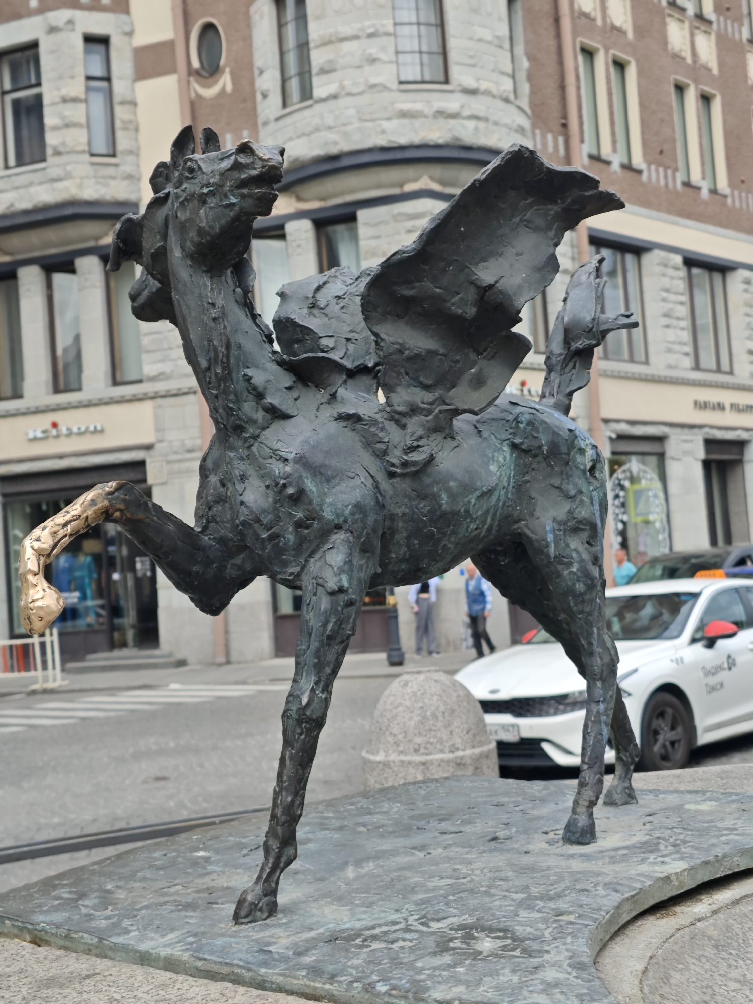 The image depicts a bronze sculpture of a winged horse with a golden hoof. The sculpture is positioned on a stone base in a public urban setting. In the background, a multi-story building with architectural details and windows is visible. Street activity is evident, with a white car, identified by Russian text and branding as a taxi, partially visible. Two individuals are walking in the background. The scene appears to be a street in Saint Petersburg, Russia, given the taxi branding and architectural style. The sculpture itself is the primary subject, with its weathered bronze texture and the striking contrast of the golden hoof.