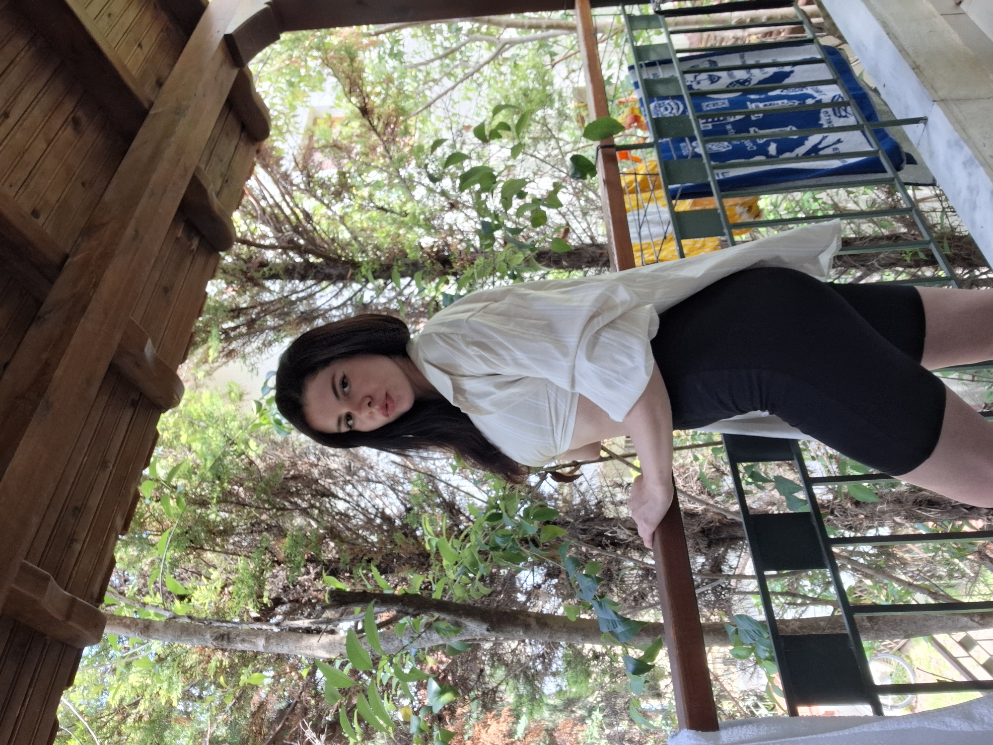 A woman stands on a balcony with a wooden roof and railing. She is wearing a white cropped shirt and black shorts. She is leaning on the wooden railing with her left hand and her right hand is resting on the railing as well. In the background, there are trees and a glimpse of buildings. Blue and white towels are hanging on a rack on the balcony.