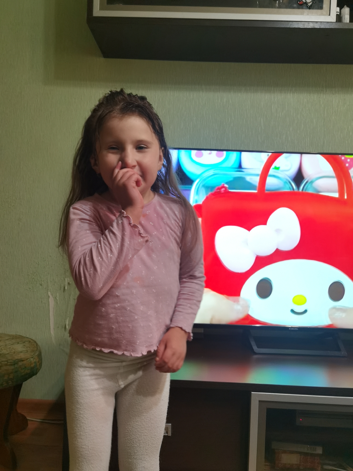 The image depicts a young, light-skinned girl standing indoors, within what is identified as the city of Chaikovsky, Russia. The girl, positioned slightly left of center, possesses long, light brown hair, which appears textured around her forehead, possibly due to dampness or styling. She is wearing a long-sleeved, light pink shirt adorned with small, scattered white or silver dots, and a ruffled cuff is visible on her right wrist. Her lower attire consists of light-colored, textured leggings, likely white or light grey. She presents a broad smile, with her right hand partially covering her mouth.
The background includes a pale green wall on the left, exhibiting visible wear, texture, or areas of peeling paint. A dark green, patterned stool or ottoman is partially visible in the bottom-left corner. Dominating the right side of the frame is a flat-screen television displaying a vibrant animated image. The screen features the character My Melody, presented as a red bag or container with a large white bow and a white face with black oval eyes, a yellow nose, and a small black smile. Other colorful, cartoon-like objects are visible in the background on the television screen. The brand name "xiaomi" is faintly visible on the lower bezel of the television.
Below the television is a dark brown media console or cabinet. The television is supported by a black stand on this console. The media console features a lower compartment on the right side, with a silver-framed panel and a visible red indicator light on an electronic device stored within. Above the television, a dark, wall-mounted shelf is present, and above that, a lighter-colored shelf holds a framed object and a small white cylindrical item.
