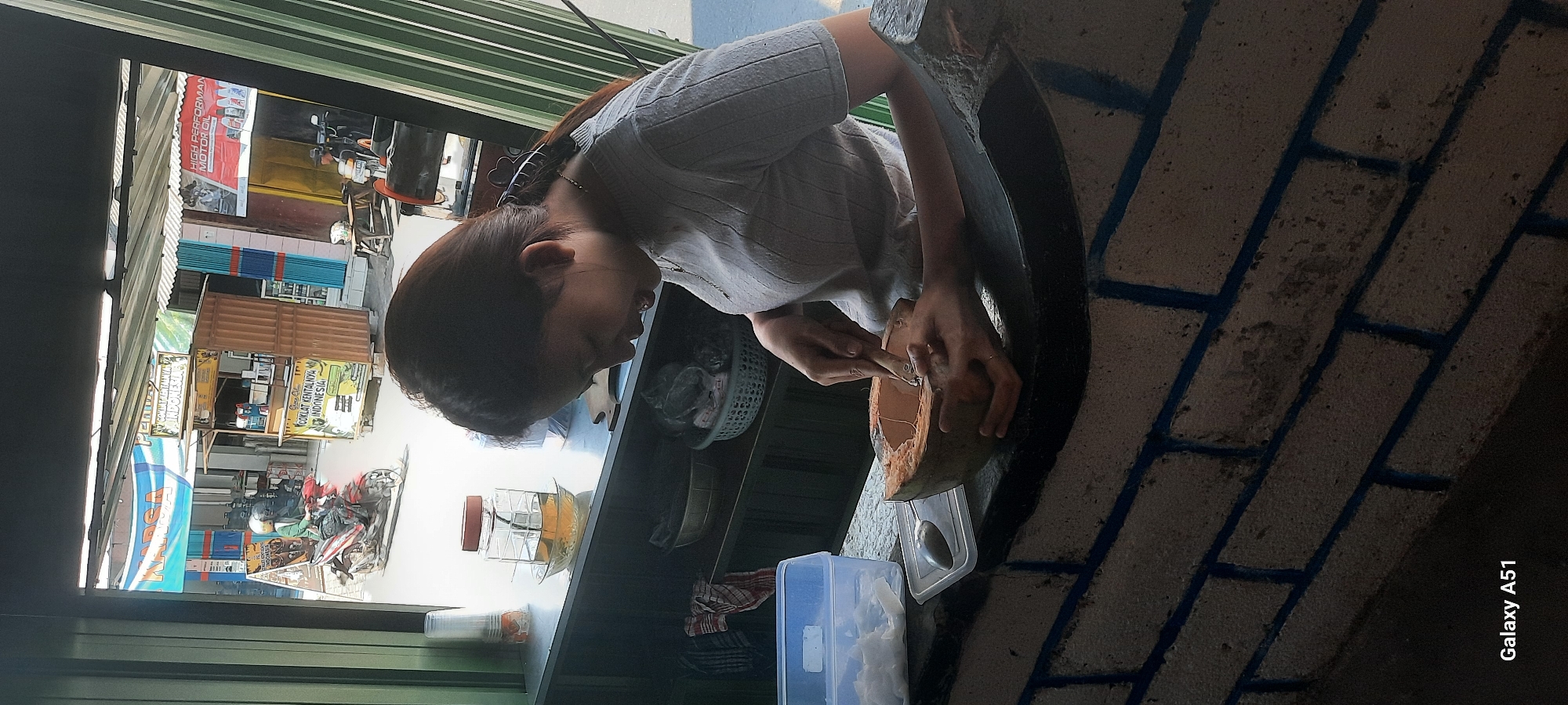 In Selopuro, Indonesia, a young woman is seen from the chest up, in profile, focused on scraping the inside of a halved coconut. She is wearing a light gray, short-sleeved shirt with a ribbed texture and a delicate necklace. Her dark hair is pulled back, revealing her face as she smiles slightly while engaged in the task. To her left, on the counter, is a blue plastic container with what appears to be shredded coconut inside, and a silver spoon resting on a white lid. Further back on the counter, a glass jar filled with amber-colored liquid, possibly tea or juice, sits on a saucer. The background reveals a street scene visible through an open window, with buildings, a food stall selling "COKLAT KENTANGNYA INDONESIA," and a person riding a motorcycle. The counter she is working on is made of dark stone, and below it, a brick wall painted with blue lines is visible. The overall lighting suggests it is daytime.
