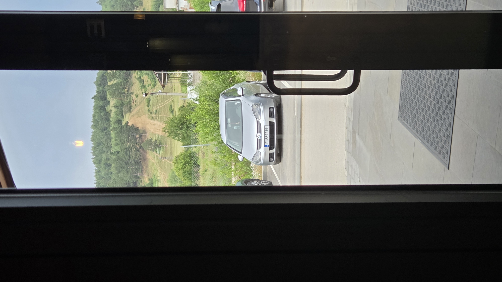 A full outdoor shot through a dark doorway shows a silver Volkswagen Golf parked in a lot, with a wooded hill in the background. A dirt path winds up the hill, with some sparse trees and power lines visible. In the sky above the hill, there is a bright, translucent orange object that resembles a lantern. The car has a European license plate that reads "E 8892 HH." The doorway has a dark frame and a dark door is partially ajar, revealing a grey tiled floor with a dark mat in front of it. The lighting suggests it is daytime, possibly overcast.