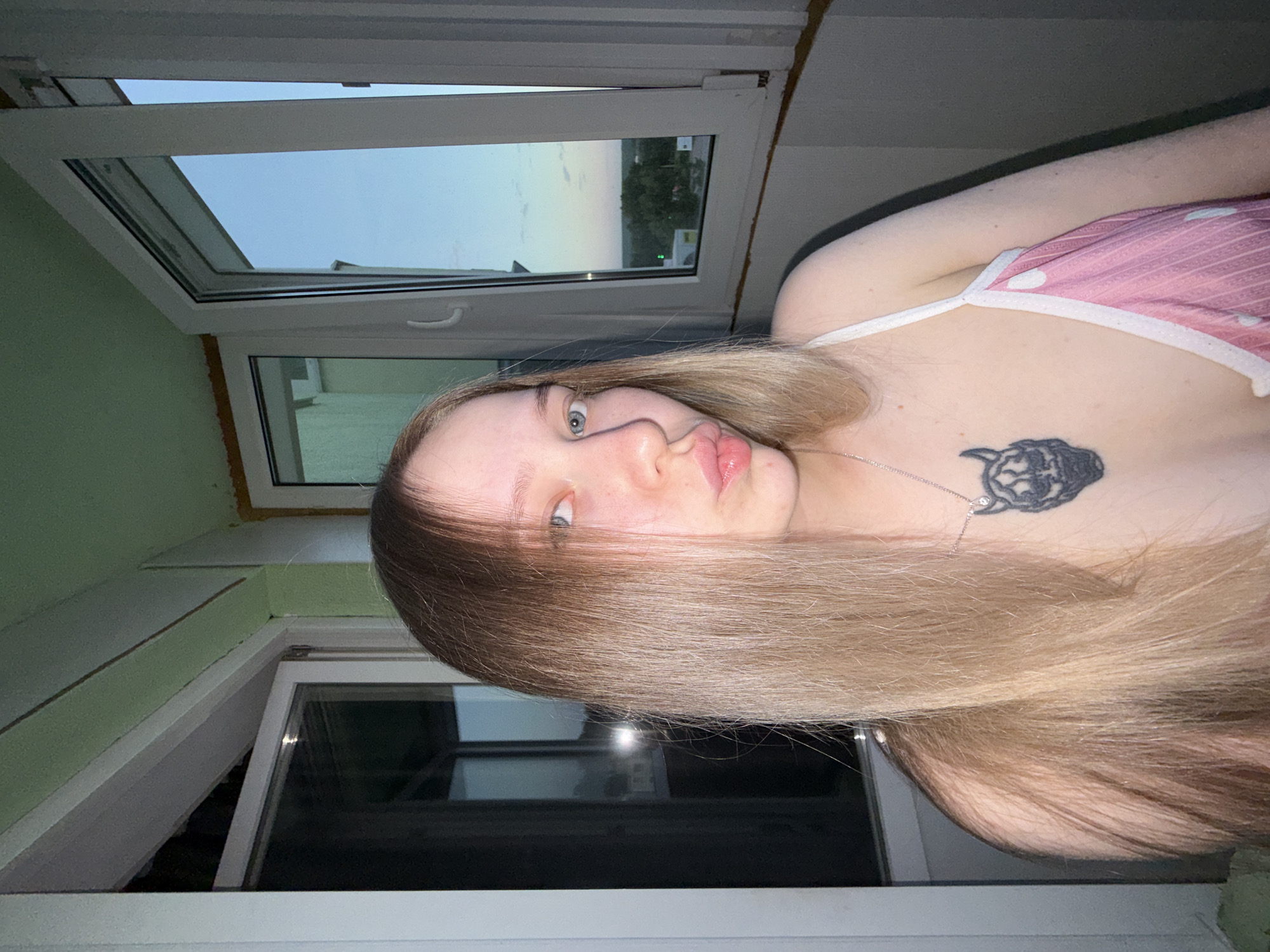 A fair-skinned person with long blonde hair partially obscuring their face is photographed indoors. They are wearing a pink and white polka-dot top with thin straps. A black tattoo is visible on their upper chest. The background features a window with a view of a landscape under a pale sky. The person is positioned in a corner of a room with light green walls. The photo appears to be taken inside a room in Chaikovsky, Russia, evidenced by visual cues in the scenery visible through the window.