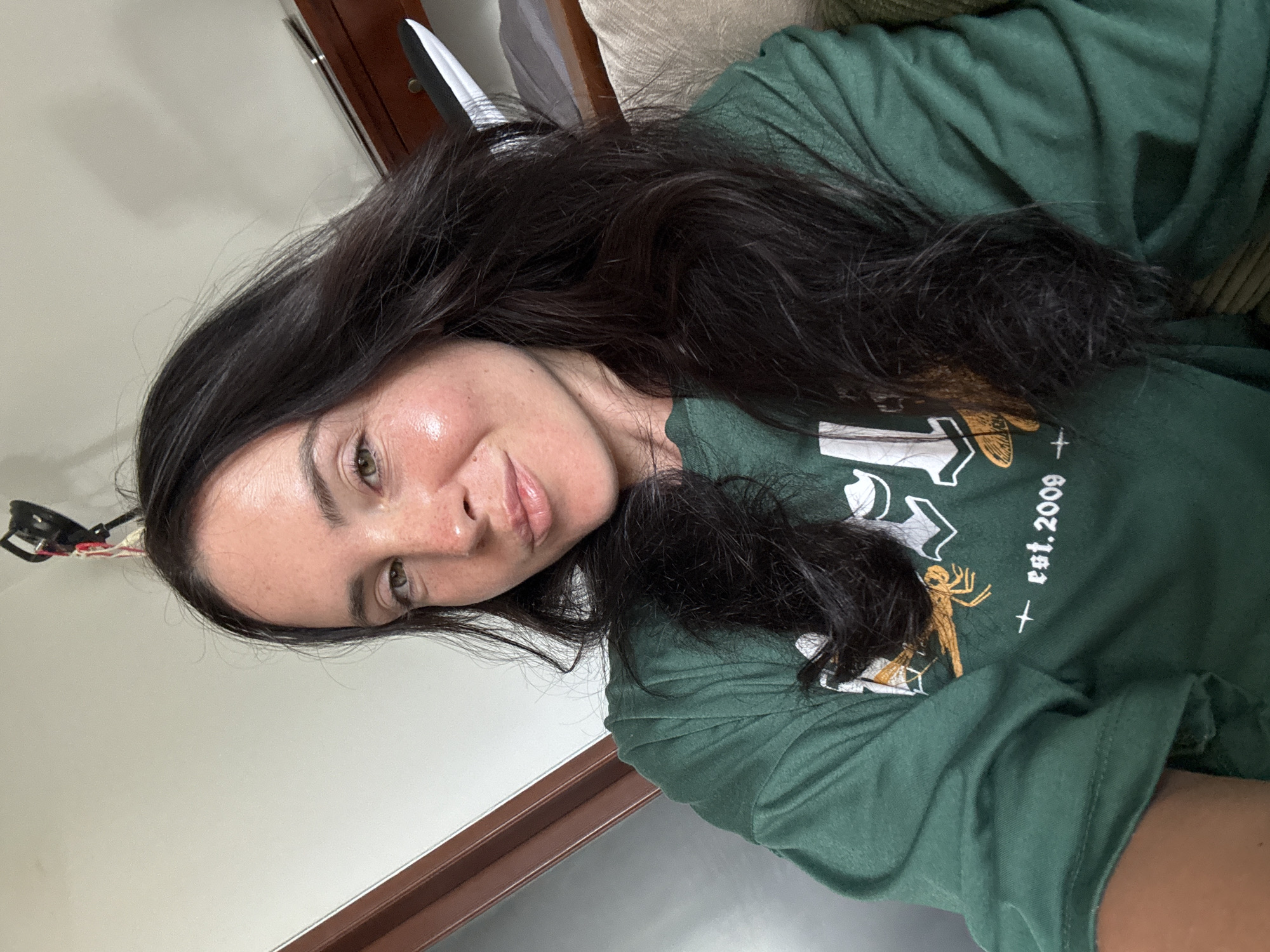 A close-up, selfie-style shot shows a woman with dark brown hair and fair skin. She is wearing a green t-shirt with a graphic design that includes the text "est. 2009" and a spider. The background is out of focus, but suggests an indoor setting with a wall, a wooden piece of furniture, and a bed. The lighting appears to be natural, possibly from a window. The image is taken from a high angle.
