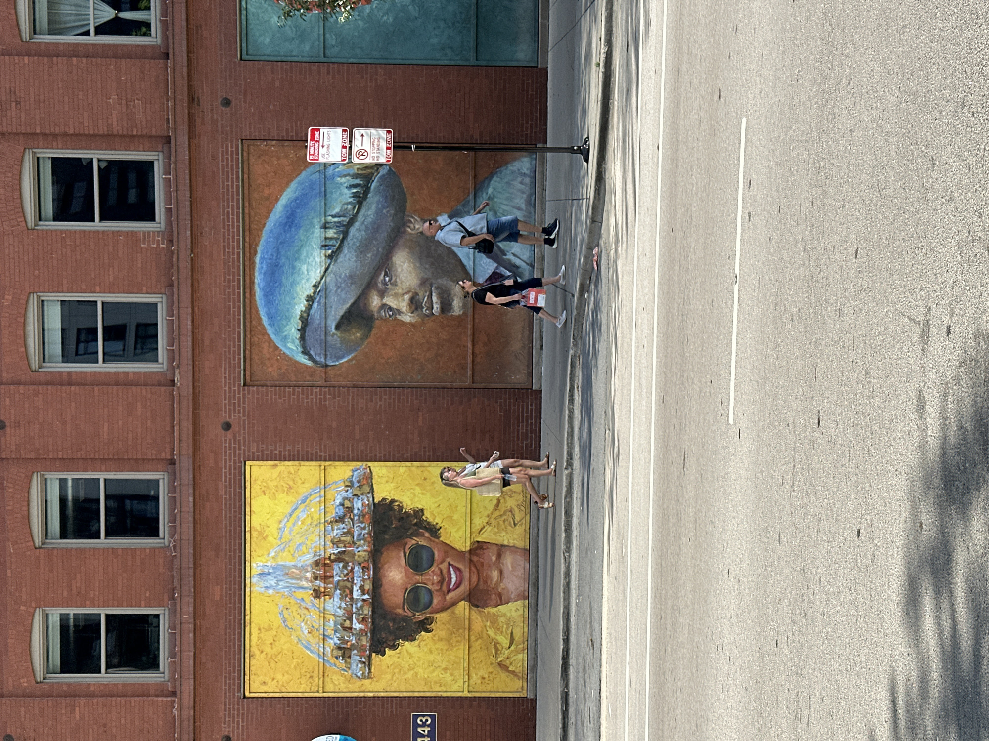 On a brick building in Chicago, Illinois, two large murals are visible. The mural on the left depicts a woman's face with dark, curly hair and large sunglasses, her head adorned with a golden crown resembling a fountain. She wears a yellow jacket and smiles warmly. Two women walk on the sidewalk in front of this mural. The mural on the right is a portrait of a man wearing a wide-brimmed blue hat, which reflects a cityscape. A couple walks on the sidewalk in front of this mural, with a pole featuring parking signs situated between them and the mural. The street in front of the sidewalk is paved with asphalt and marked with white lines. The buildings have multiple windows and are made of red brick. The lighting suggests a sunny day.