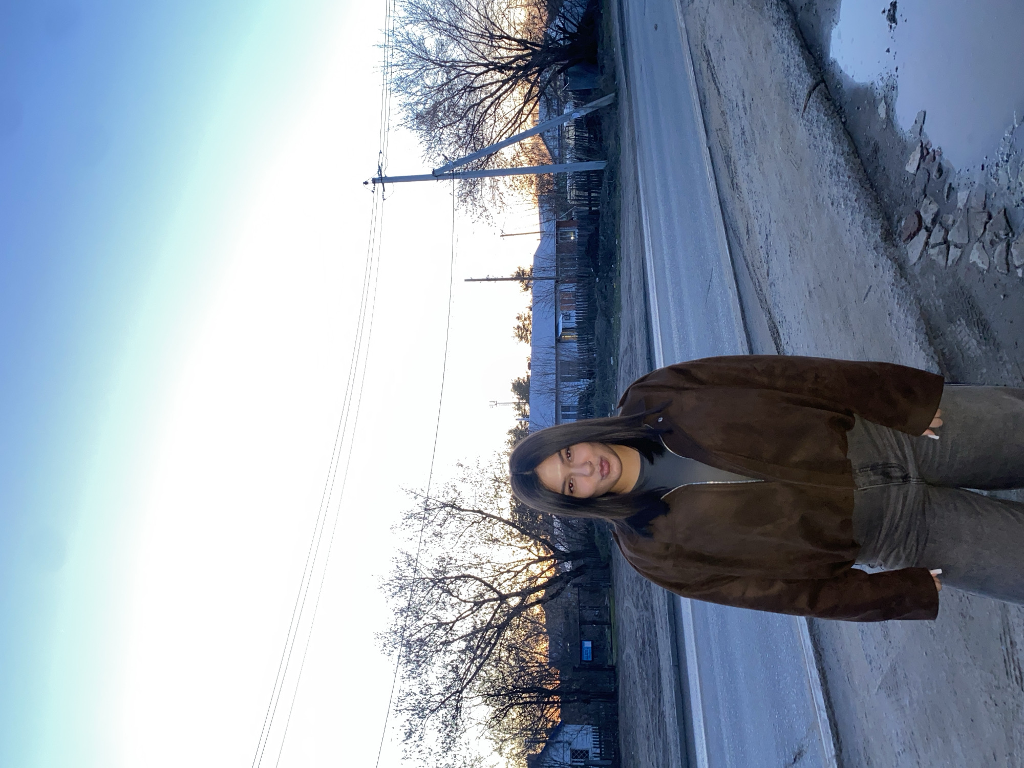 A woman stands on the side of a street. She is wearing a brown jacket and gray pants. The street is wet and muddy. There is a puddle in the foreground. In the background, there are trees and houses. A power pole is visible, with lines running across the image. The sky is light blue, indicating dusk or dawn. The location is likely Chaikovsky, Russia, based on the overall aesthetic of the image.