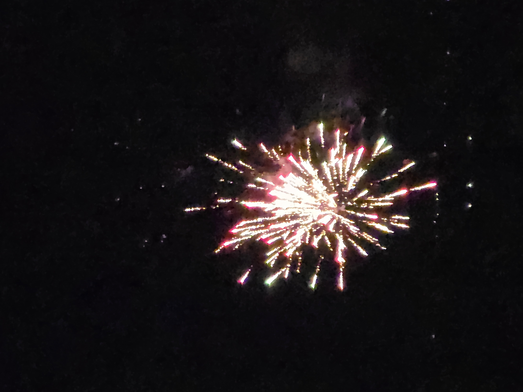 The image depicts a firework display against a dark night sky. The firework is centrally located in the frame, exhibiting an exploding burst of red, gold, and white streaks of light emanating from a central point. There are a few scattered, fainter stars visible in the background. The location is Unknown, Unknown.