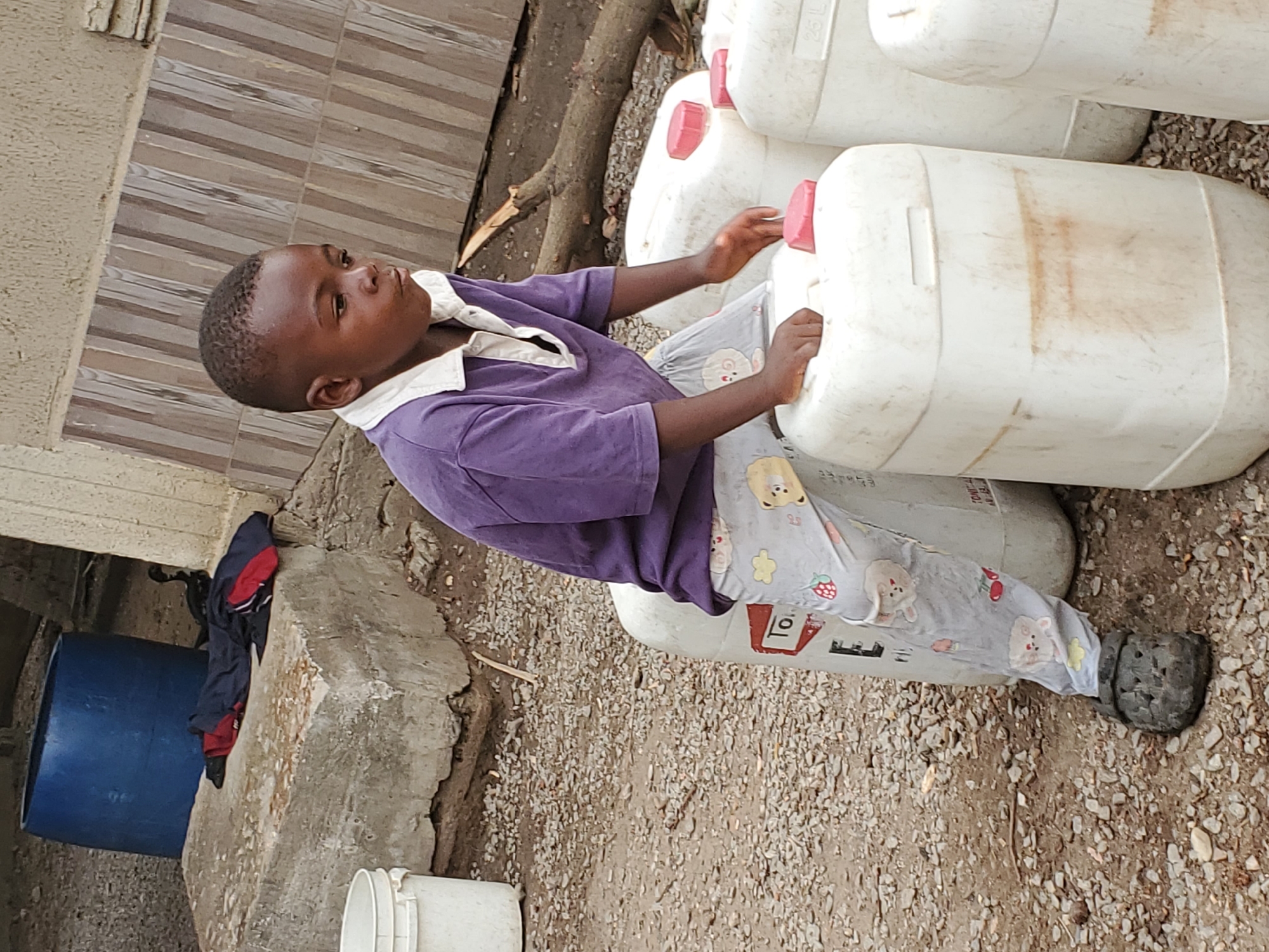 An outdoor, eye-level shot captures a young Black boy interacting with several white plastic jerrycans. The boy, likely between 4 and 7 years old, is dressed in a short-sleeved purple polo shirt with a white collar and light grey pajama bottoms adorned with small cartoon bunnies, strawberries, and yellow flowers. He wears dark, possibly rubber, clogs.
He stands beside a stack of large, off-white plastic containers, each fitted with a red screw-on cap. The containers show signs of use, with some dirt and reddish-brown stains. The boy holds onto two of these containers with both hands, looking slightly upwards and to his right with a neutral, focused expression.
The ground beneath him and extending into the midground is composed of small, light-colored gravel and dirt. In the background, to the left, a section of a building or wall with horizontal brown and grey striped tiles is visible. Below this, a large blue barrel and rough concrete blocks are present. A dark blue and red jacket or piece of clothing hangs over one of the concrete blocks. Natural daylight illuminates the scene, suggesting it is daytime. On one of the containers, the text "25 L" is visible, indicating its capacity, and a partial word "To" can be seen on a label on another. The setting depicts an everyday scene in an outdoor, possibly domestic or storage area in Mararaba, Nigeria.