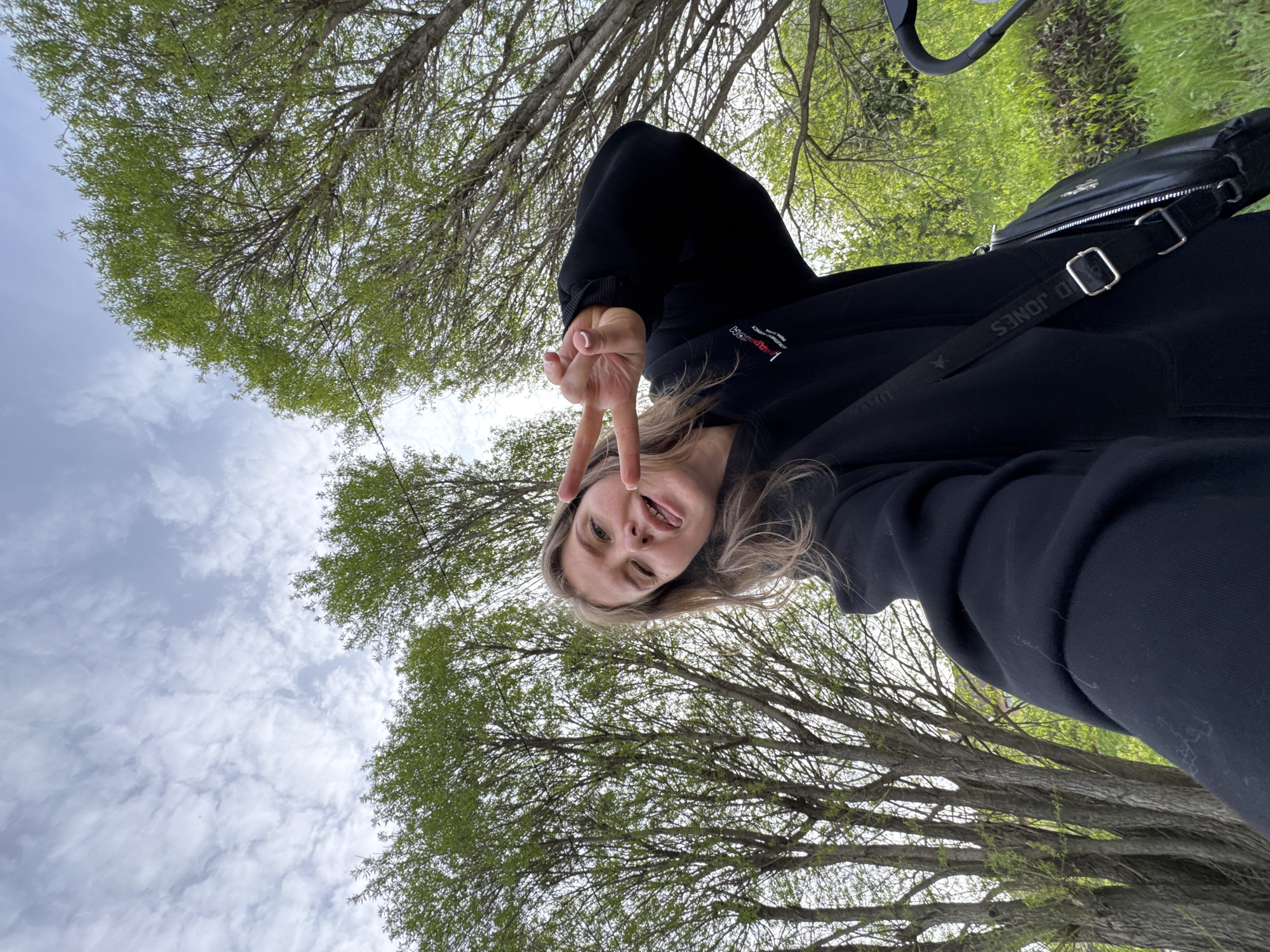 A woman is depicted from a low-angle perspective, likely a selfie. She is wearing a black hoodie and making a peace sign with her hand, sticking her tongue out, and winking. She also has a black bag. The background features trees with green foliage and a cloudy sky. The photo was taken in Горный, Russia.