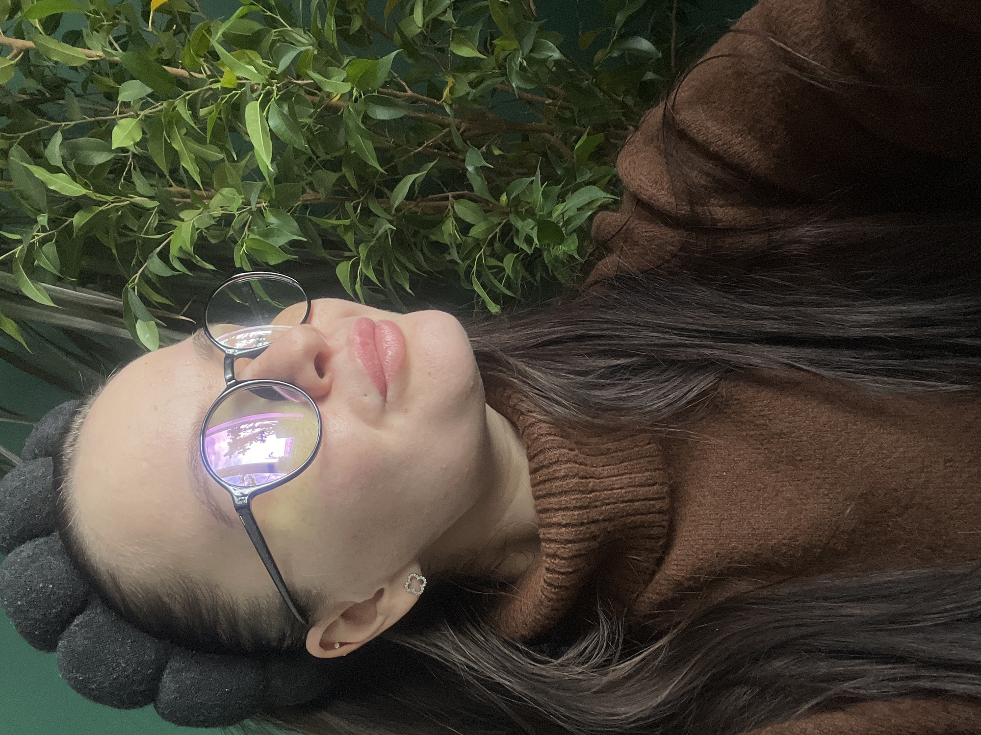 A person wearing glasses and a brown sweater lies on their back with a plant behind them. The setting appears to be indoors. The person has dark hair and is wearing earrings shaped like flowers. There is a black flower-like object near their head. The location is Kazan, Russia.