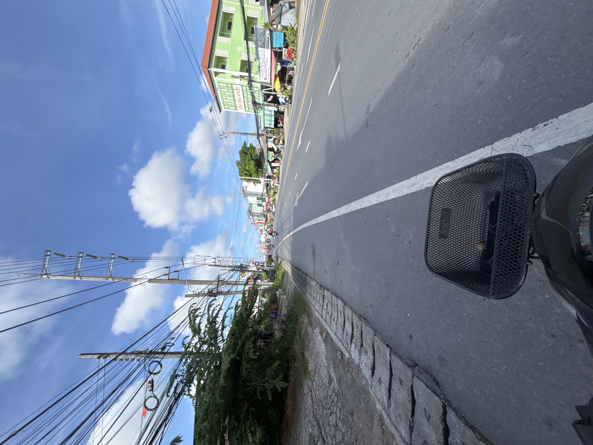 The image captures a view from a bicycle basket, looking forward along a street in Chalong, Thailand. The road is paved with asphalt and marked with white lane dividers, indicating multiple lanes of traffic. On the left side of the road, there is a sidewalk area with a concrete drainage channel. Beyond the sidewalk, there are trees and utility poles with a complex network of electrical wires overhead. On the right side of the road, several buildings are visible, appearing to be shops or residences with green exteriors and red roofs. Some of these buildings have awnings and signs in what appears to be Thai script. There are several motorbikes on the road, some moving and some parked. The sky is blue with scattered white clouds. The overall scene depicts a typical streetscape in a Southeast Asian city, likely during daylight hours given the brightness and clear sky.