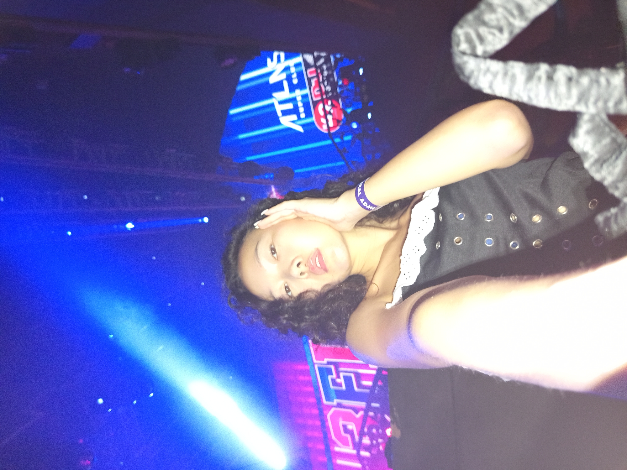 A young woman with dark curly hair and an open mouth is taking a selfie in a dimly lit venue with blue and pink lighting. She is wearing a black corset-style top with a white lace collar and a purple wristband. Her left hand is resting on her cheek. In the background, there are bright lights and signs, one of which appears to say "ATLAS SUPER CLUB" with red accents. The setting appears to be a nightclub or entertainment venue.