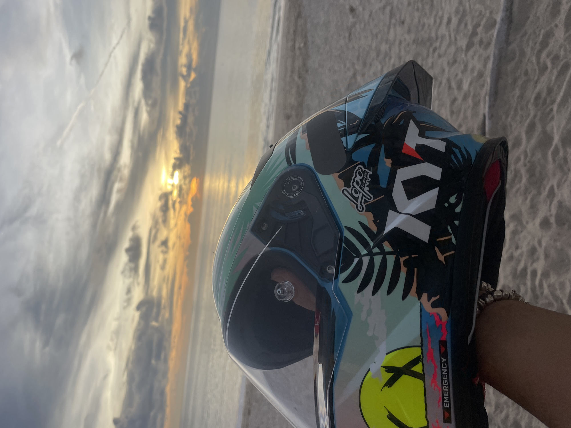 Here's a detailed description of the image:
**Location:** The image appears to be taken on a beach in Choeng Thale, Thailand, as evidenced by the beach, water, and the setting sun, suggestive of a coastal location.
**Subject:** The primary subject is a motorcycle helmet with a colorful design. The helmet features a mix of tropical imagery (palm leaves) with graphic elements like a yellow smiley face and bold lettering and logos.
**Foreground:** The helmet is held up in front of the sunset. There is a person's hand visible holding the helmet. The person is wearing a bracelet.
**Background:** The background consists of a beach scene with water and a dramatic sky with setting sun and clouds. The sun is reflecting on the water surface. The sand appears to be light colored.
**Details:** The helmet has a clear visor. The side of the helmet has the word "EMERGENCY" and there is a logo on the upper portion of the helmet.
**Overall Impression:** The image captures a moment of leisure or travel, possibly related to motorcycling, set against the backdrop of a scenic tropical beach at sunset.