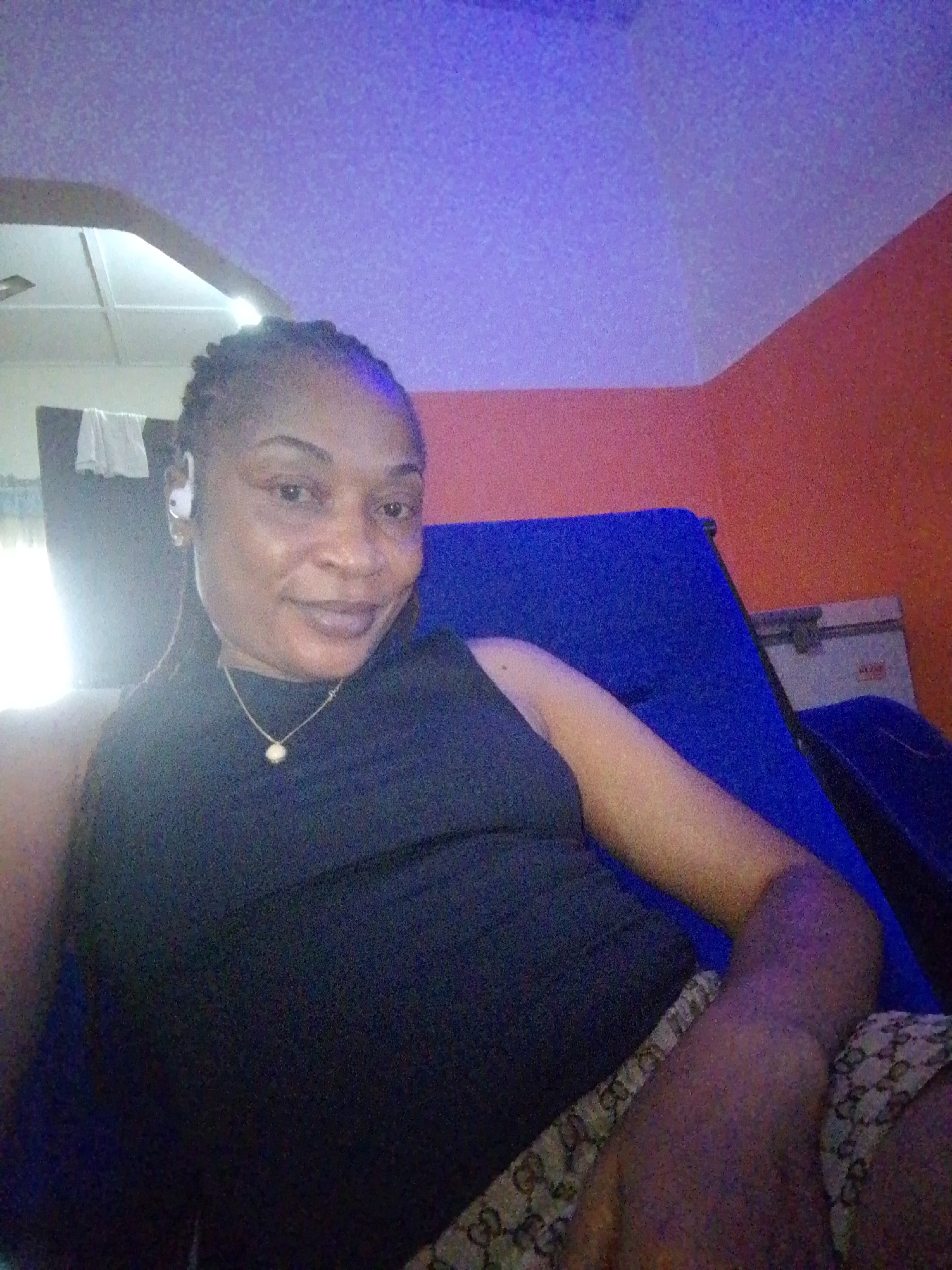 A person with braided hair and earrings is sitting on a blue chair in a room. The person is wearing a black top and patterned shorts. There is a white ceiling with lights and an orange wall in the background.