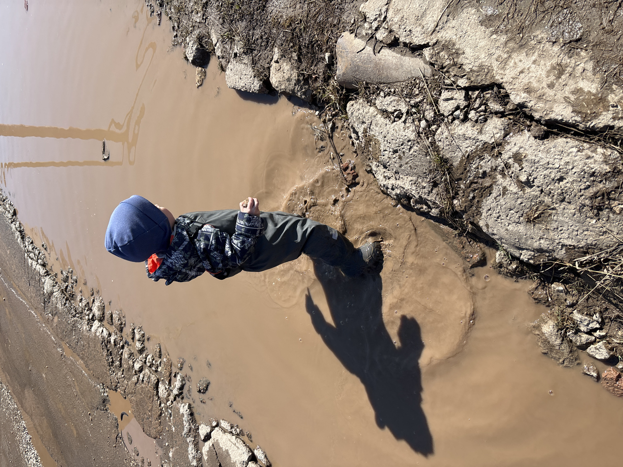 A child stands in a large, rectangular, brown puddle in Ferma, Russia. The child wears a blue beanie, a patterned jacket, dark pants, and dark shoes. The puddle is surrounded by broken concrete and dirt on all sides. Part of a road is visible in the lower left corner. The child's shadow is cast on the right side of the puddle.