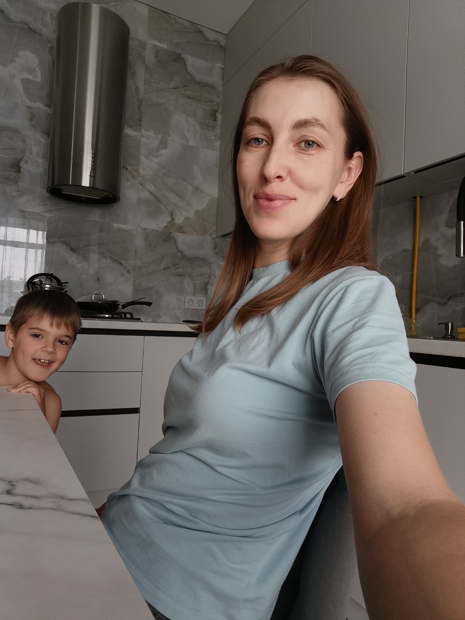 In this medium shot, a woman and a young boy are in a modern kitchen in Chaikovsky, Russia. The woman, positioned in the foreground and taking the selfie, has shoulder-length brown hair and is looking directly at the camera with a slight smile. She is wearing a light blue t-shirt. The boy is to her left, peeking from behind a marble-patterned countertop. He has short brown hair and a wide, happy smile, looking towards the camera with his left arm resting on the counter.
The kitchen features sleek, light-colored cabinetry and a marble-patterned backsplash behind the stove area. A stainless steel range hood is prominently mounted on the wall above the stove, which has cookware on it. The overall aesthetic is clean and contemporary. The lighting suggests it is daytime, with natural light possibly coming from a window that is partially visible in the background. The mood appears relaxed and cheerful, with both subjects smiling.