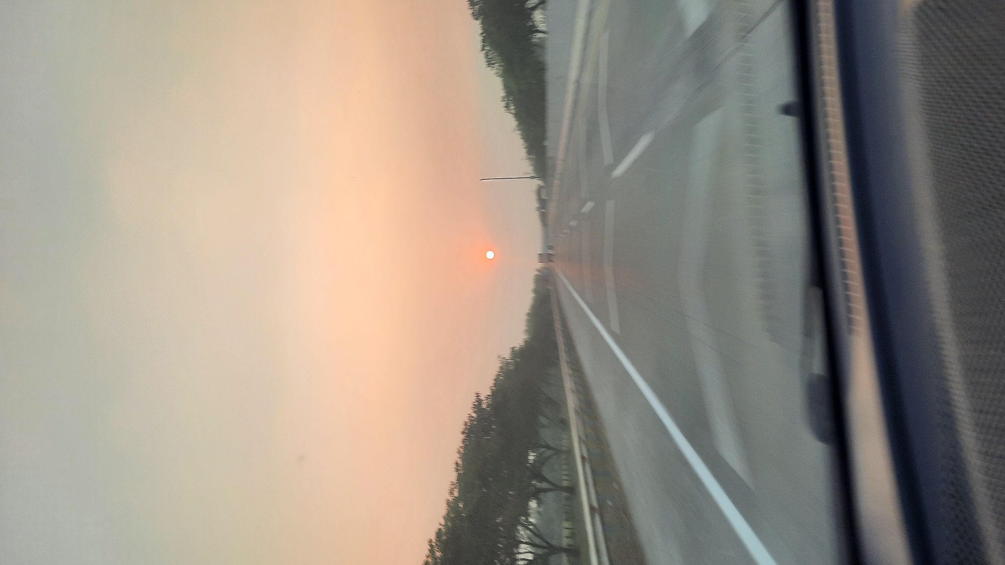 The image depicts a sunset view from a vehicle traveling on a highway, possibly the Ngawi-Kertosono toll road in Ngawi, Indonesia. The sun is a small, bright orange orb positioned centrally in the hazy sky. The sky transitions from a pale yellow-orange at the horizon to a lighter pastel shade towards the top. Trees line both sides of the highway, appearing as a dark, blurred silhouette due to motion. The road itself is asphalt, with clear white lane markings visible. In the distance, a few vehicles can be seen on the highway. The foreground shows a portion of the vehicle's interior, including the dashboard, which is blurred and out of focus. The overall impression is one of movement and the tranquil beauty of a sunset on a long journey.