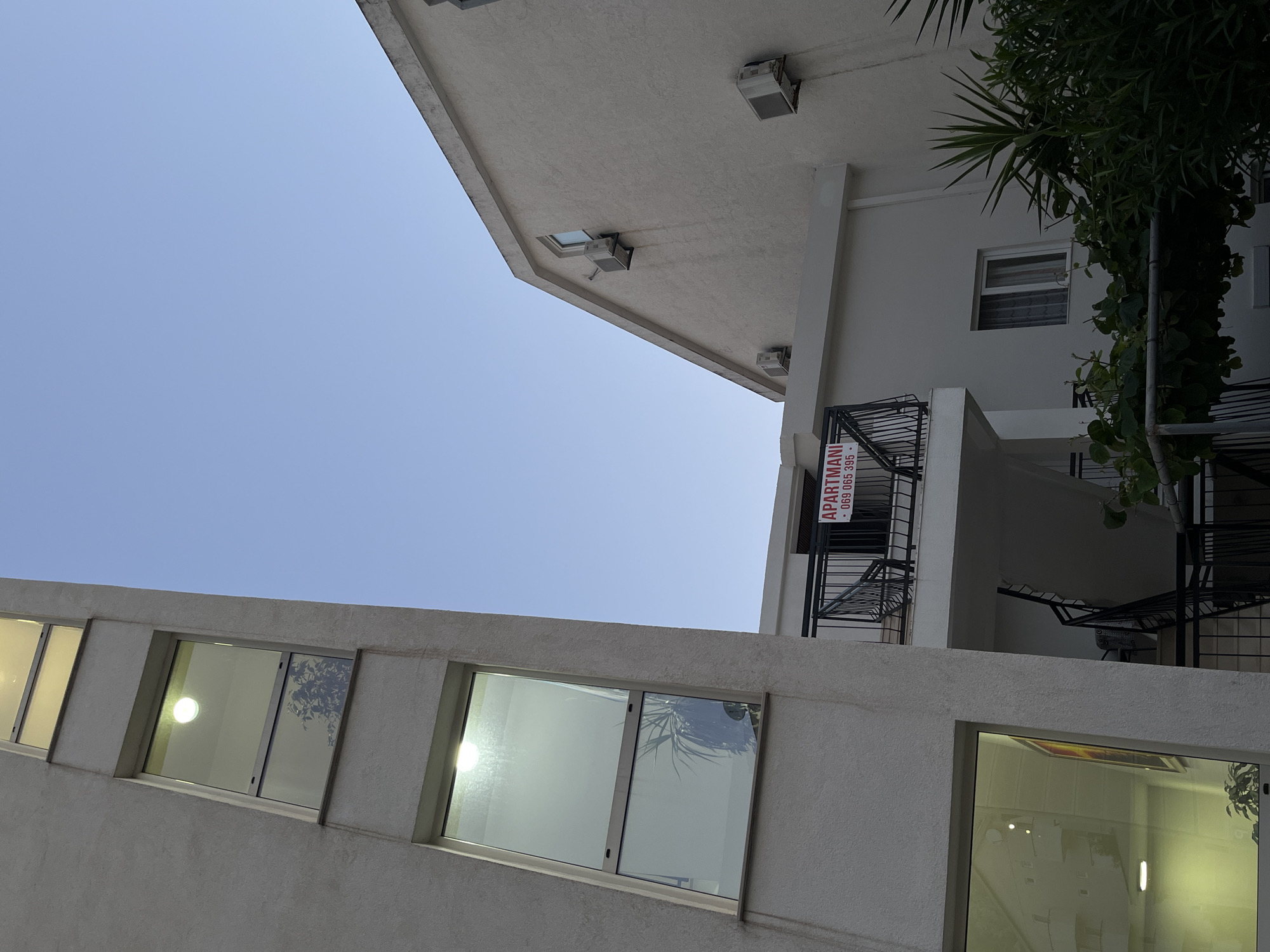 This is a low-angle outdoor photograph of an apartment building in Budva, Montenegro, under a clear blue sky. The building's facade is textured and painted a light color. On the left, a section of the building with multiple rectangular windows extends upwards. The windows have a reflective quality, showing interiors with artificial lighting and some reflections of foliage and possibly other buildings.
To the right of this section, another part of the building is visible, featuring a peaked roofline. Two air conditioning units are mounted on the exterior wall of this section. Below the peaked roof, a balcony area is visible with a black metal railing. A red and white sign is attached to the railing, reading "APARTMANI" followed by a phone number.
In front of the building, there is a dark metal staircase leading upwards to the balcony. Lush green foliage, including palm-like plants and vines, is growing around the base of the staircase and in the foreground, obscuring parts of the lower levels of the building. The overall impression is of a residential or tourist accommodation building, possibly during the day or early evening.