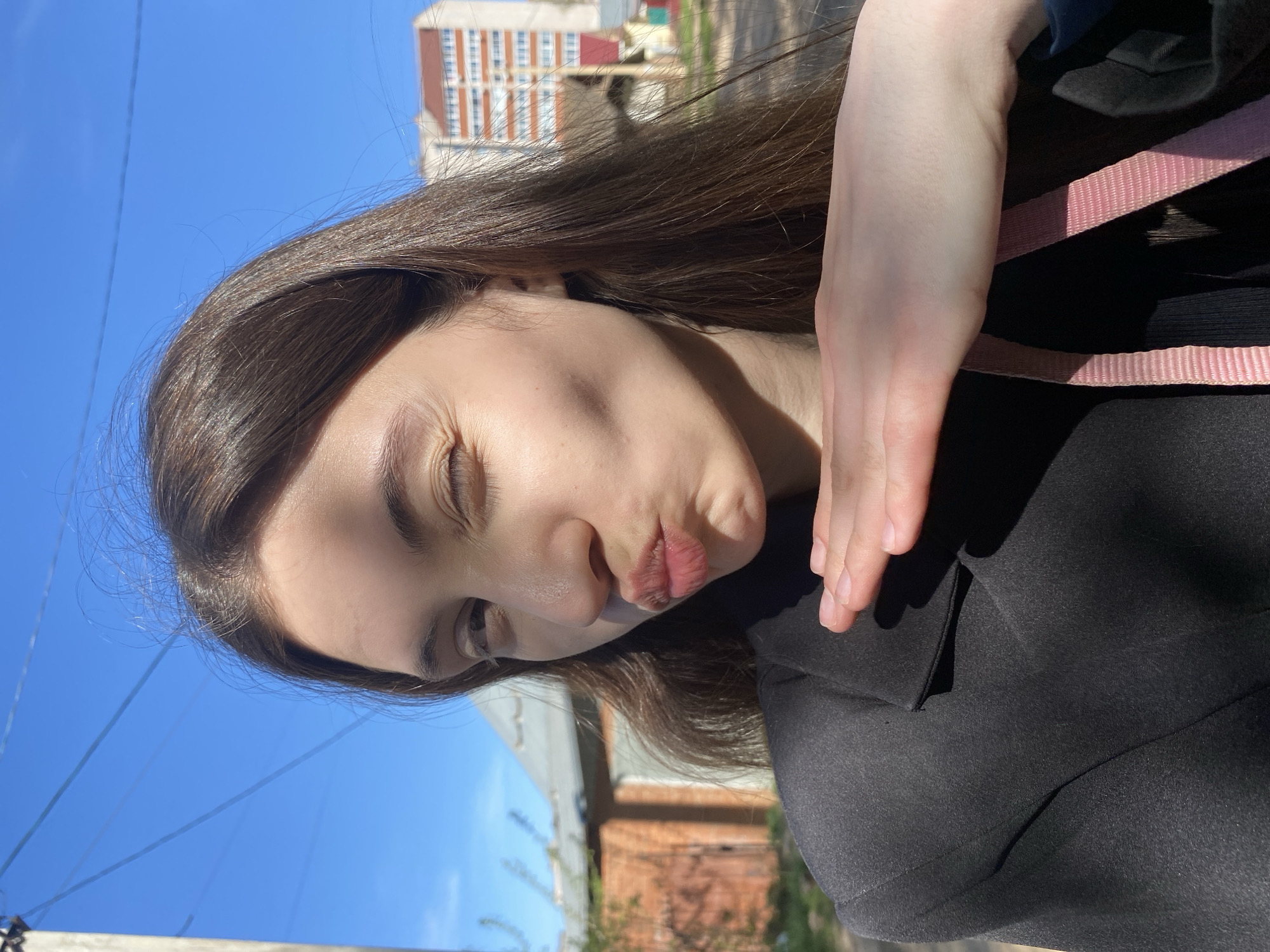 A young woman is seen in an outdoor selfie-style photograph. She has long, brown hair and is wearing a black top with a pink strap. Her hand is near her face, and she has a slight pout. The background includes a blue sky, buildings, and some greenery.
The specific location is not discernible as the architecture is generic.