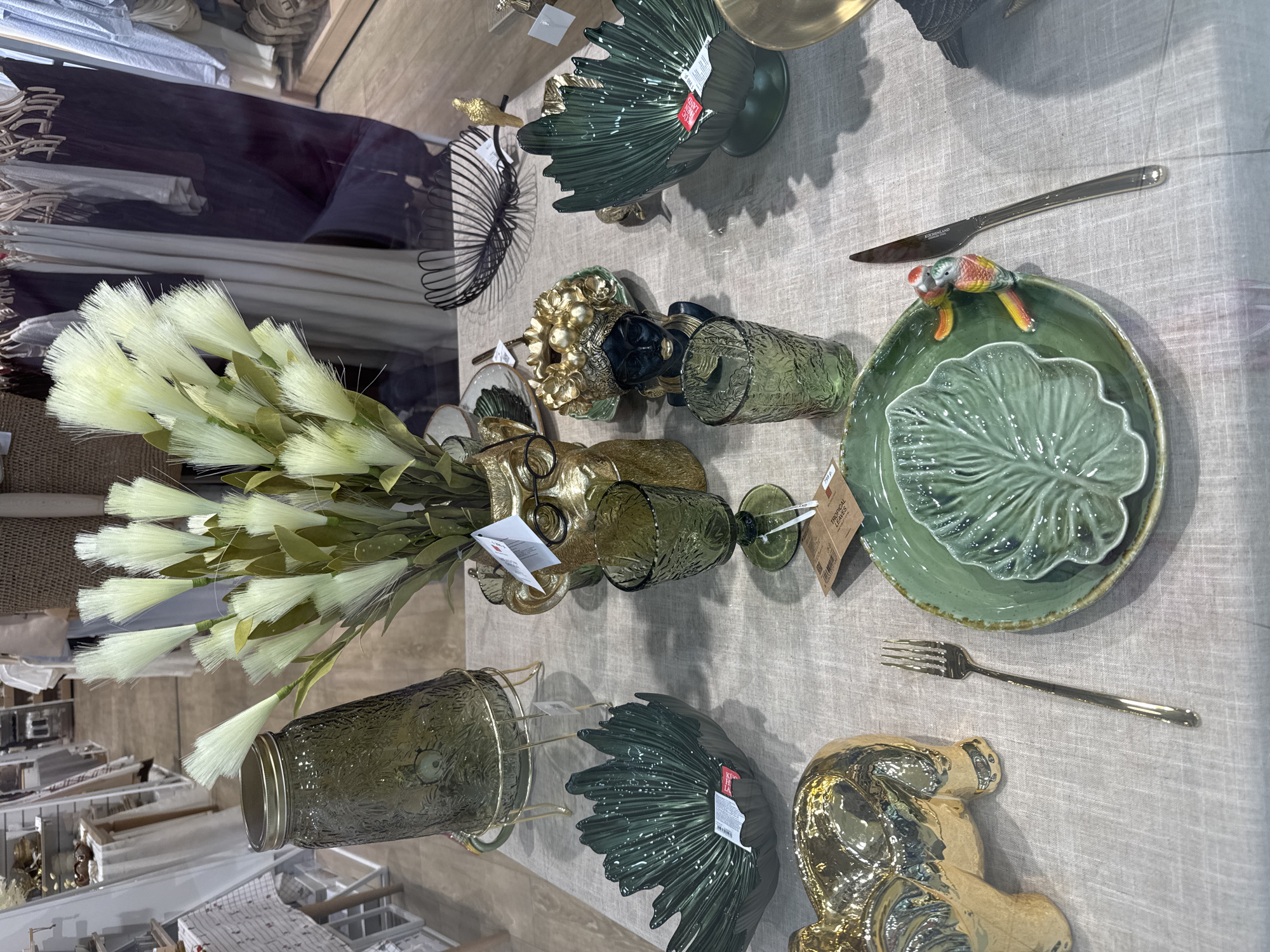 This image captures a vibrant display of home decor and tableware, likely set up in a retail store or showroom. The scene is viewed from an elevated perspective, looking down onto a table covered with a light-colored, possibly linen, textured fabric.
The primary subjects are various decorative objects and functional tableware, predominantly in shades of green and gold. Key items include several green glass vases and tumblers, some with textured patterns. A large bouquet of light yellow-green, feathery artificial flowers resembling pampas grass is prominently displayed in a green vase.
The tableware features green ceramic plates, with some designed to resemble tropical leaves, and one plate has a small, colorful ceramic parrot figurine perched on its rim. Gold-colored cutlery, specifically a knife and a fork, are laid beside the plates. Other decorative elements include various gold-colored figurines, one appearing to be a stylized animal, and another resembling abstract golden fruit. A dark green, shell-patterned bowl and a black wire basket with a spiraling design also adorn the table.
The background shows reflections of other merchandise and shelving in a glass partition or display window, indicating an indoor commercial setting. There is no human activity taking place; the scene is a static product arrangement.
Notable details include the cohesive green and gold color palette, which creates an elegant and nature-inspired aesthetic. The textures of the glass, ceramics, and fabric add visual depth. Visible text includes "KÜCHENLAND" on the knife, "TROPICAL LEAVES" on a price tag, and "799 p." (presumably indicating a price in Russian Rubles, consistent with the Saint Petersburg context) along with "KIWI" on another tag. The time of day is not discernible, but the lighting is even and artificial.