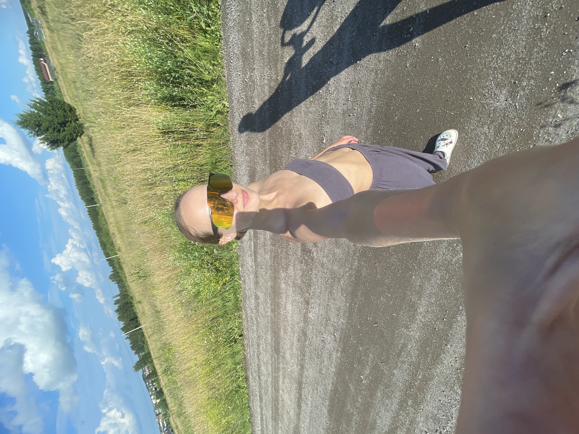In a selfie taken outdoors on a sunny day in the village of Podshivalovo, Russia, a young woman with blonde hair pulled back and wearing oversized mirrored sunglasses, a dark grey bandeau top, and dark grey loose-fitting pants poses for the camera. Her right arm is extended, holding the camera. She is standing on a gravel road, with a grassy, slightly overgrown field behind her that slopes uphill. The field is dotted with various grasses and wildflowers and bordered by a line of trees. In the distance, houses and more trees are visible on the horizon under a blue sky with white, puffy clouds. The woman's shadow stretches across the road and into the field. She is wearing white sneakers with pink trim.