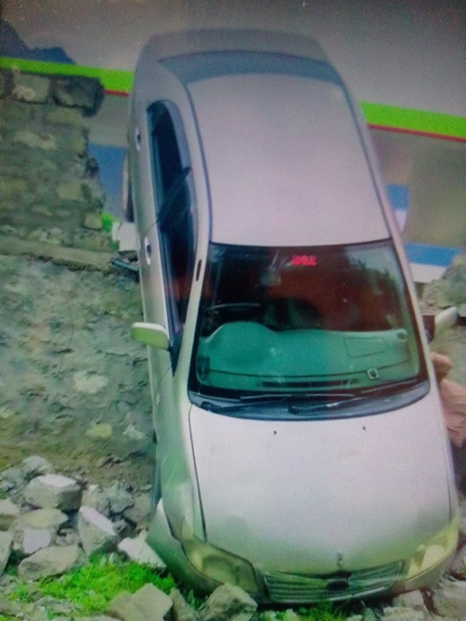 A silver sedan car is depicted at an angle, appearing to be partially off a road or path and stuck in a ditch or on uneven terrain. The front driver's side of the car is embedded among rocks and dirt, with patches of green grass visible around the front bumper.
In the background to the left, there is a stone or concrete wall. Beyond this wall and above it, a blurry mountainous or hilly landscape can be seen. A structure with distinct green, blue, and red striped sections is also visible in the background, behind and to the right of the wall. The lighting suggests it is daytime.
The car is a four-door sedan, likely an older model. The windshield has a slight green tint, and an indiscernible red object or reflection is visible inside the upper part of the windshield. The front grille and headlights are visible, but there is no clear branding or text. A side mirror is extended on the driver's side. On the far right edge of the main image, a person's arm or hand is partially visible near the front passenger side of the car, suggesting someone might be inspecting the vehicle or its predicament. The overall image quality is low, with noticeable grain and blurriness.