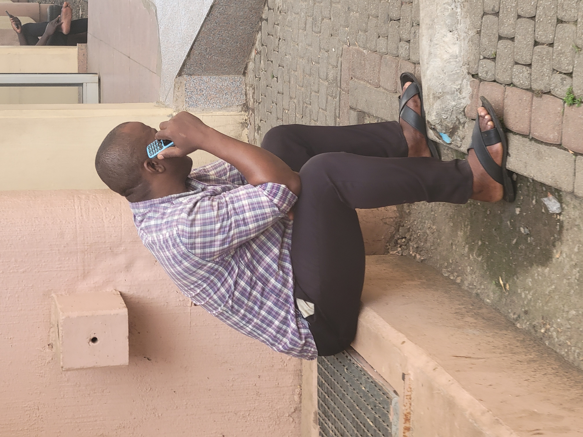 The image, rotated 90 degrees counter-clockwise, depicts a man resting on a light-colored concrete ledge or steps, engrossed in a phone call. The man, with dark skin and short or bald hair, is wearing a purple and white plaid short-sleeved shirt, dark trousers, and black leather sandals. He holds a light blue keypad phone to his right ear, his left hand resting casually.
In the upper portion of the image, to the left and slightly behind the main subject, another person's legs and bare feet are visible, also resting on similar light-colored steps. This individual wears dark trousers, and one hand, holding a dark object, can be seen.
The setting appears to be an outdoor or semi-outdoor space in Abuja, Nigeria, featuring various architectural elements. To the right of the main subject, a section of paved ground with interlocking grey and reddish-brown blocks is visible, interspersed with small patches of green vegetation. A concrete curb separates this paved area from the ledge. Below the man's legs, a metal grate, possibly part of a drainage system, is embedded in the ground. The surrounding walls and structures are primarily light brown or tan.
The scene conveys a calm and relaxed atmosphere during what appears to be daytime. No specific weather conditions are indicated. There is no visible text on any objects or surfaces within the image.