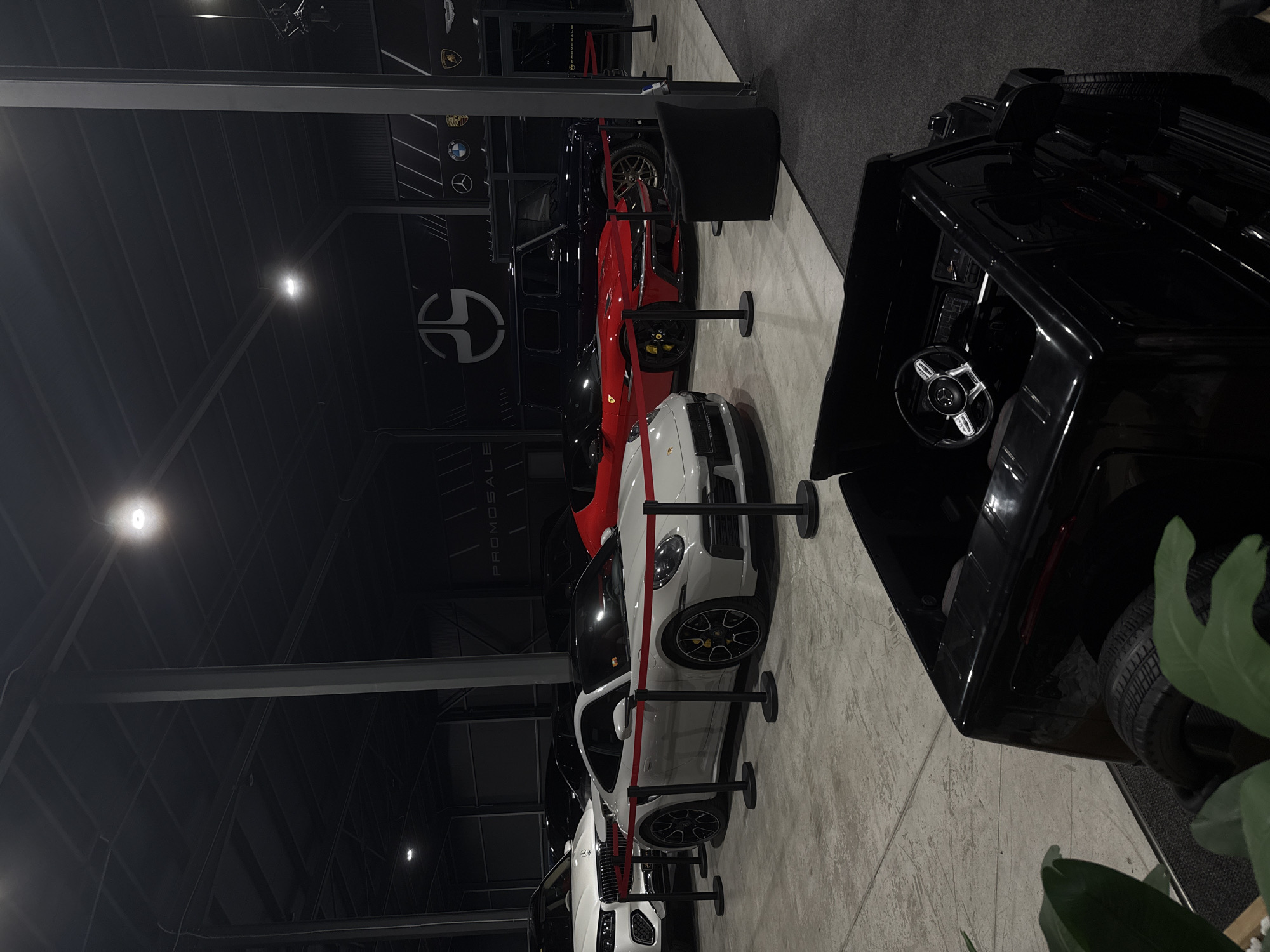 The image shows a car showroom in Plovdiv, Bulgaria, featuring a display of luxury vehicles. Several cars are visible, including a black SUV, a red Ferrari, and white Porsches, separated by stanchions. The showroom has a high ceiling with overhead lighting and metal support beams. Branding for "PROMOSALE" is visible on the wall. A black chair is positioned next to the SUV. The flooring appears to be a light gray concrete or tile. Part of a potted plant is visible in the foreground. Logos for various car brands are on the wall near the ceiling.