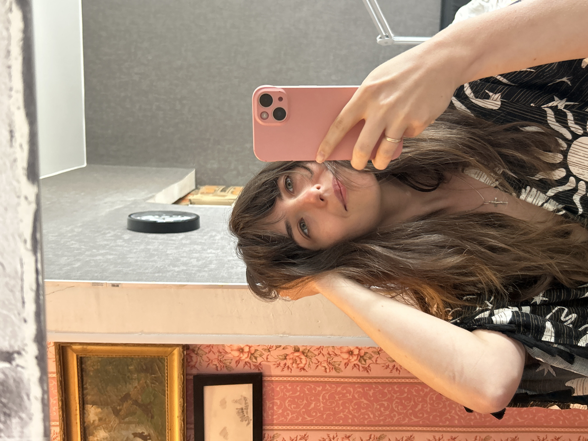 A young woman is taking a selfie in a mirror. She is holding a pink smartphone with her right hand. The reflection in the mirror shows a room with pink floral wallpaper, two framed pictures on the wall, a gray countertop, and a black circular object on the countertop. The location is suggested to be Saint Petersburg, Russia, but there is no visible evidence in the image to confirm this.