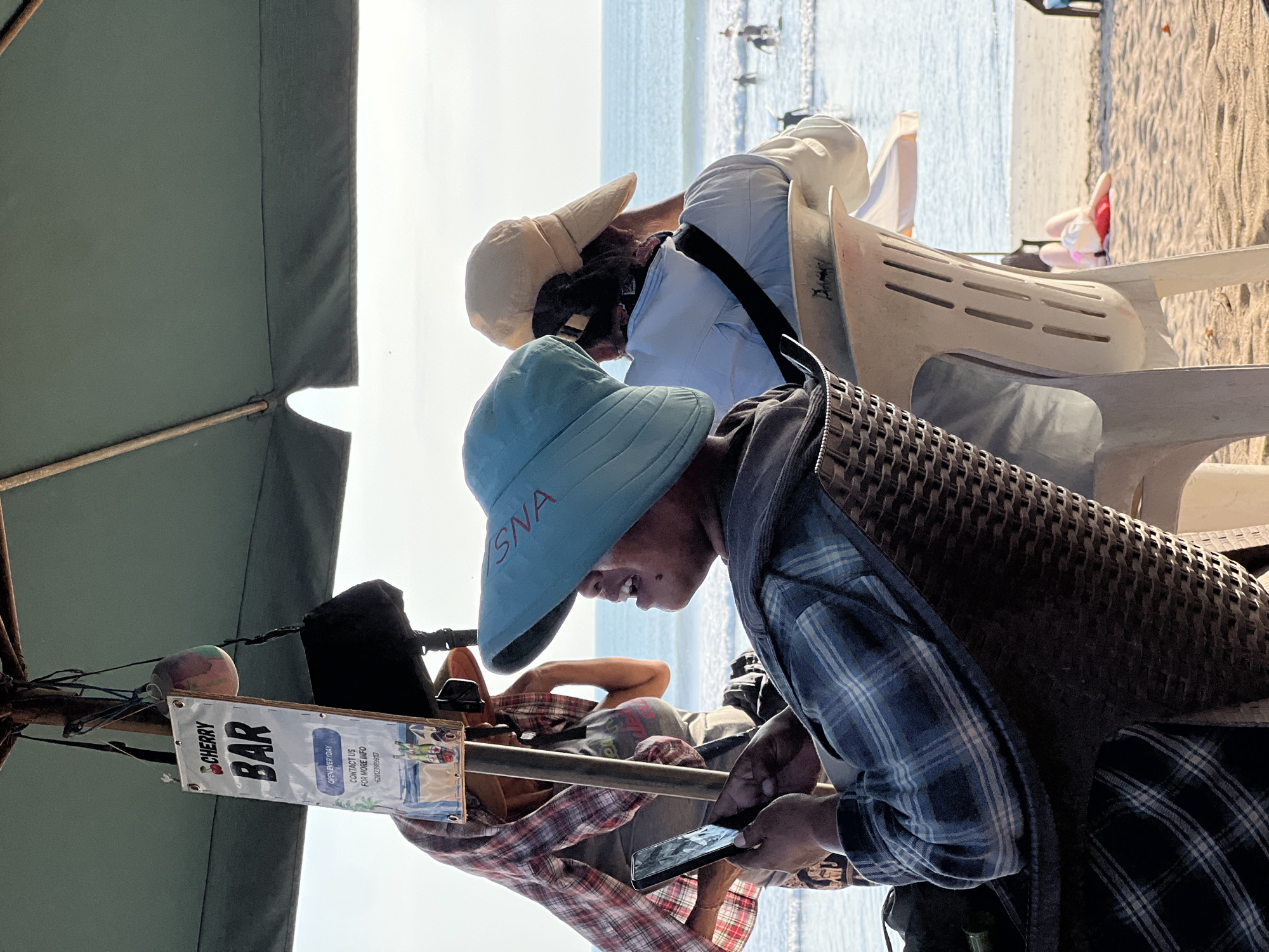 The image depicts a beach scene in Kuta, Indonesia, under a large green umbrella. In the foreground, a person wearing a blue and white plaid shirt and a light blue hat is seated in a wicker chair, looking at a smartphone. The hat has the word "SNA" written on it in red. Behind this person, another individual is visible, wearing a light blue shirt and a beige cap, facing away from the camera. A sign for "CHERRY BAR" is hanging from the umbrella's pole, indicating it is open every day and providing a contact number. The background shows the ocean with gentle waves and several people in the water, possibly surfing. A person is also lying on the beach in the distance. The overall atmosphere suggests a relaxed, coastal environment.