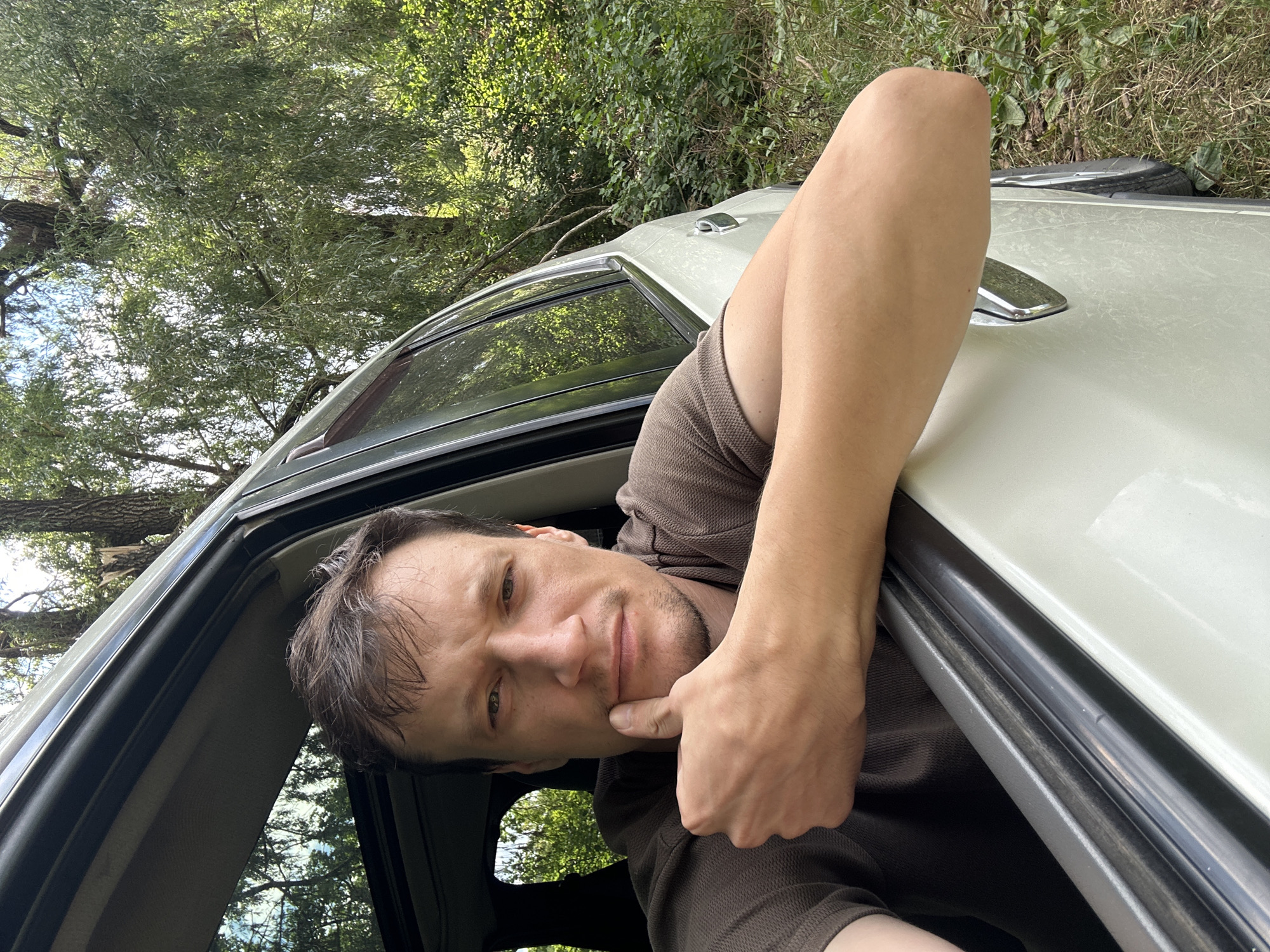 A man with short brown hair and a brown shirt is leaning out of a car window, with his right arm resting on the window frame. He is making a "thumbs up" gesture with his right hand, and his thumb is touching his chin. The car is white and appears to be in a wooded area, with trees and foliage visible in the background. The lighting suggests it is daytime. The man's facial expression is neutral, and he is looking directly at the camera. No specific location in Makiv, Ukraine can be determined from the image.