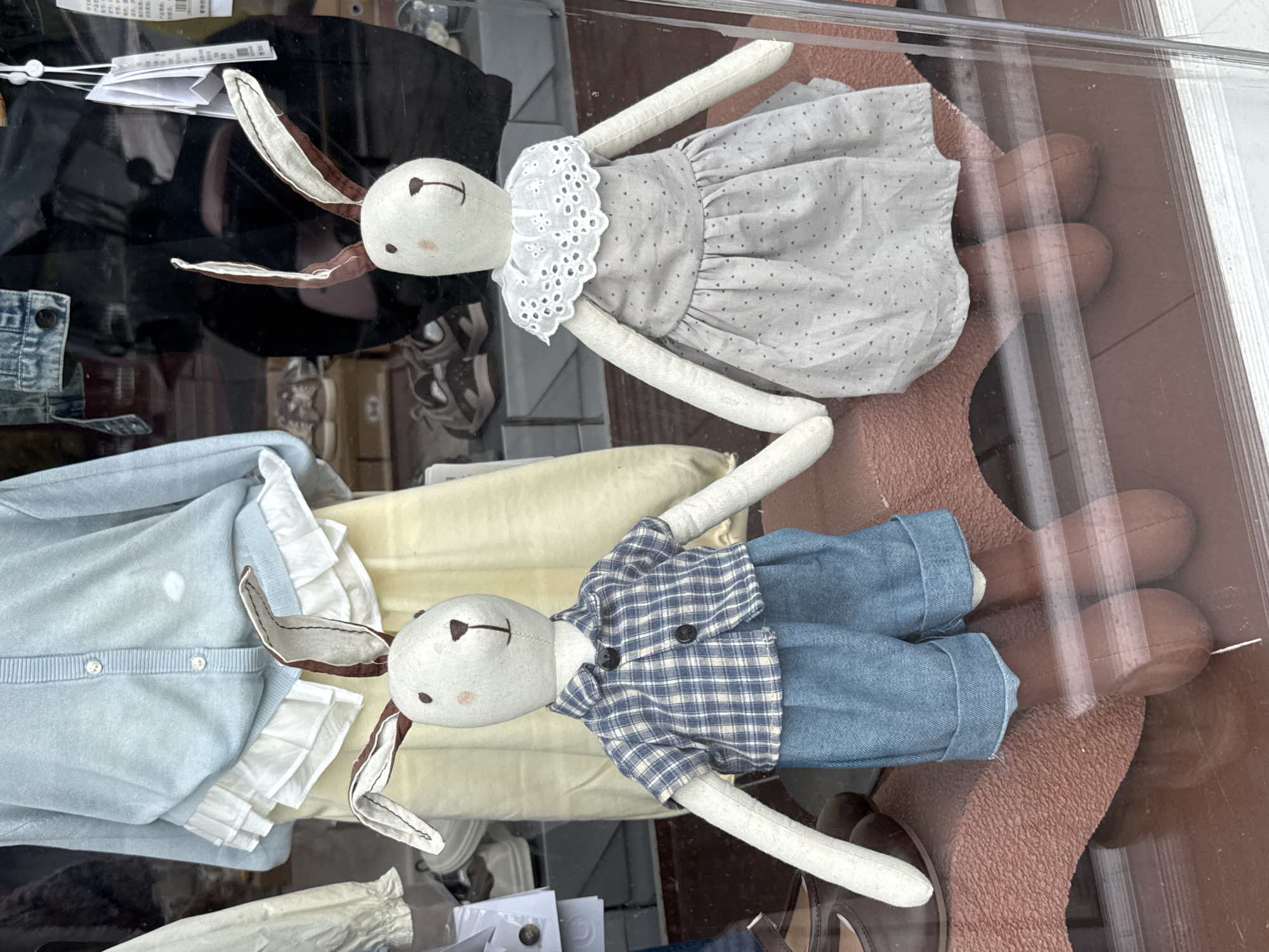 Two fabric rabbit dolls are prominently displayed in a storefront window in Wulingyuan, China. The doll on the left features a white body, brown-lined ears, embroidered black eyes, pink blush marks, and a brown embroidered nose and mouth. It wears a blue and white plaid short-sleeved shirt with dark buttons and cuffed blue denim shorts. Its feet are brown. The doll on the right mirrors the facial features and body construction of its counterpart, dressed in a light grey dotted dress with a white embroidered lace collar. Its feet are also brown. The two dolls are positioned side-by-side, with their inner hands touching, suggesting they are holding hands. They sit on a rust-colored, textured display surface. Behind them, within the store, other children's clothing items are visible, including a light blue cardigan with white ruffles over a light yellow skirt on a mannequin, and various shoes on shelves in the background. Reflections on the glass storefront create horizontal lines across the lower portion of the image.