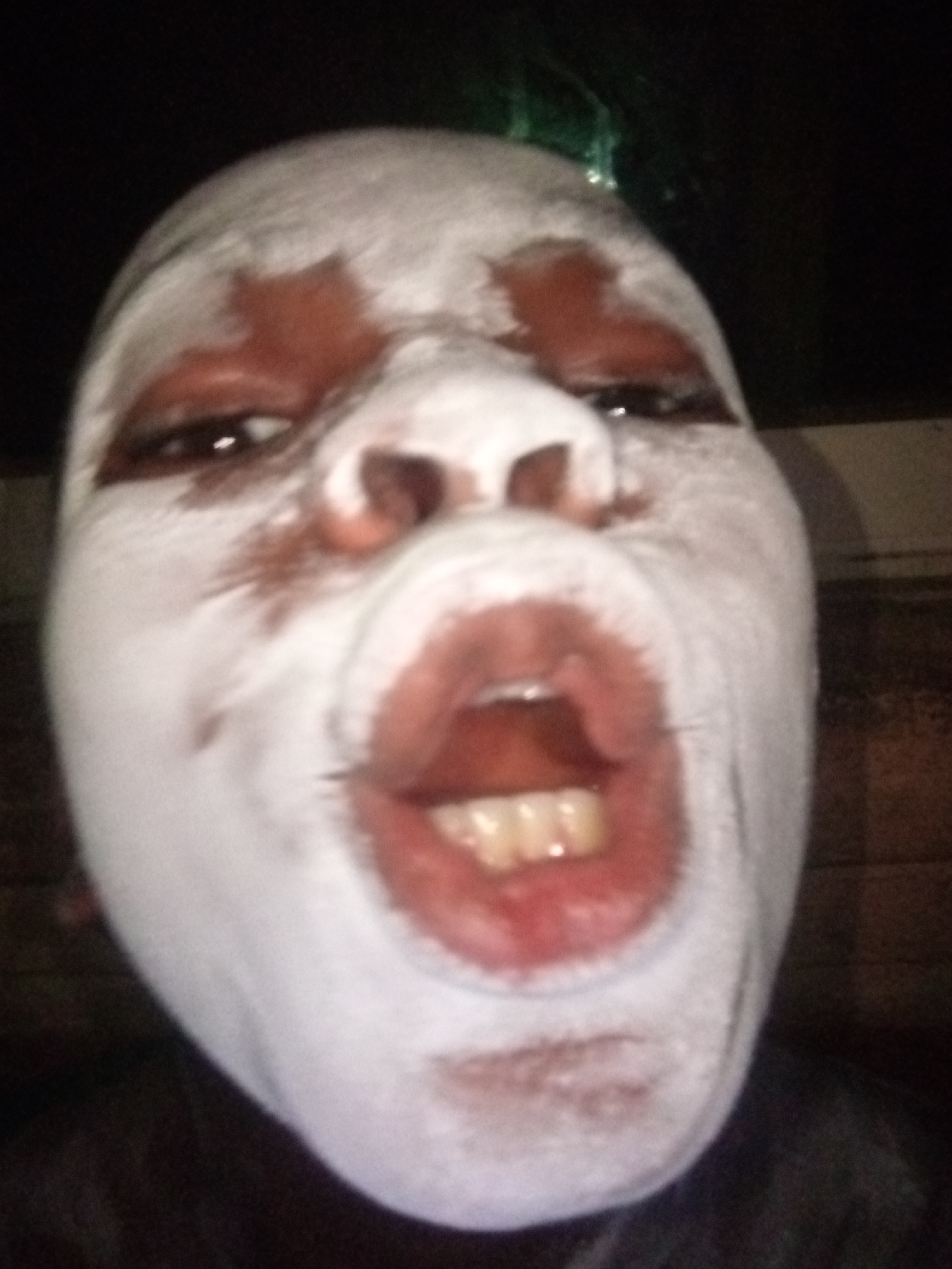 The media file presents a close-up, head-on portrait of a person whose face is extensively covered in white makeup. The individual has dark eyes that gaze directly at the viewer, and their mouth is wide open, revealing their teeth and tongue, suggesting an intense or theatrical expression, possibly mid-shout, song, or dramatic gesture.
Reddish-brown pigment is visible around the eyes and cheekbones, creating a stark contrast against the white base. A hint of green color, possibly from hair or a head accessory, can be seen at the top right edge of the frame. The background is dark and indistinct, indicating a low-light setting, likely indoors or at night. The person's skin tone is visible in areas where the white makeup is thinner or absent, particularly around the eyes and mouth. The overall appearance suggests a character or performer, possibly for a cultural event, street theater, or celebration in a location like Onitsha, Nigeria. No text is visible in the image.
