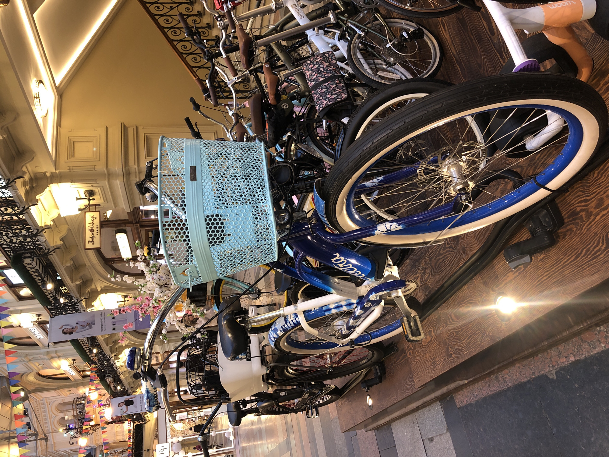 The image shows a display of bicycles, some with baskets, on a wooden floor inside the GUM shopping mall in Moscow, Russia. The background features ornate architectural details including wrought iron railings, decorative lighting, and banners advertising Huawei products. The overall environment suggests a high-end retail setting within a historic building.