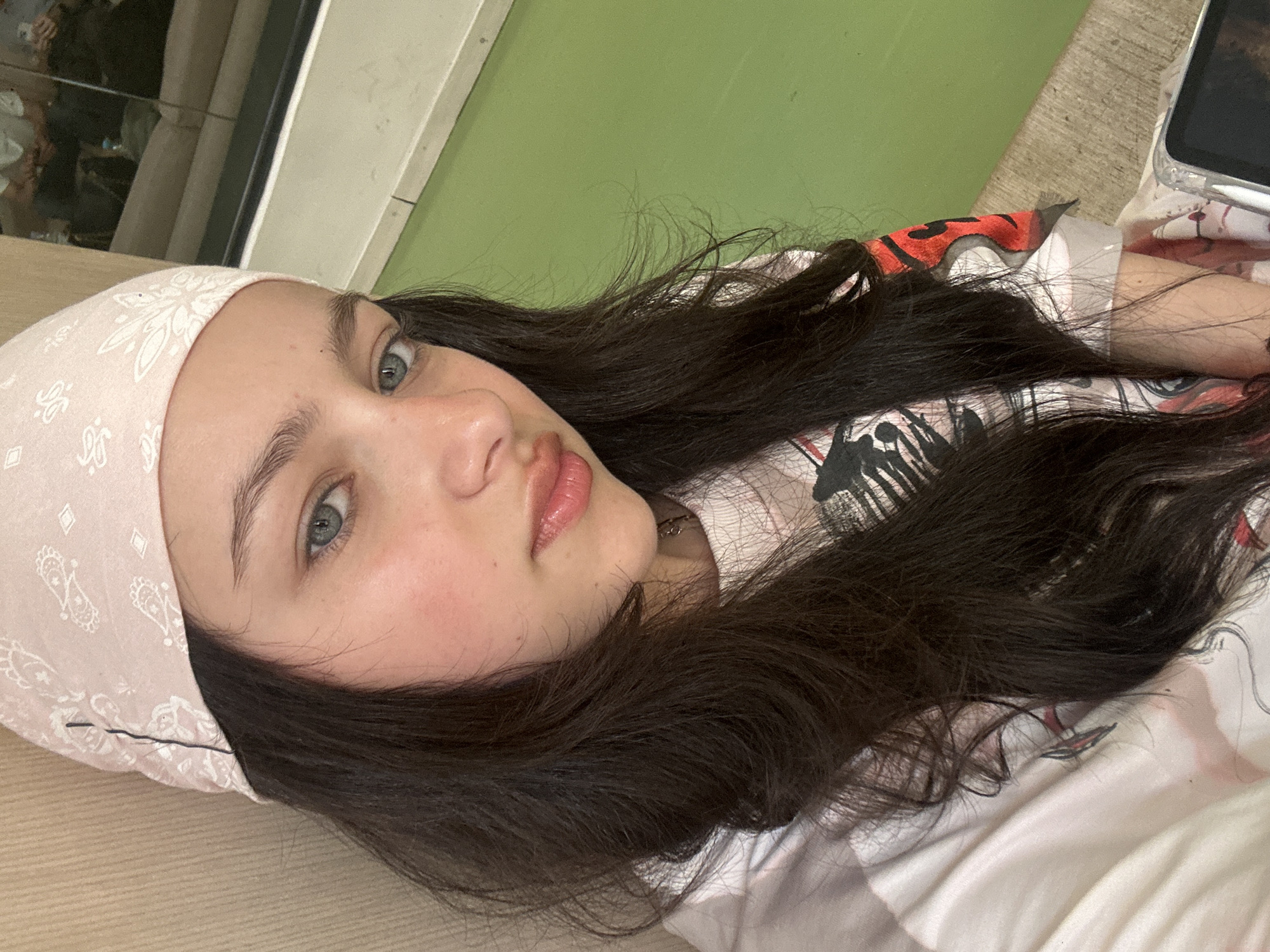 A young woman with fair skin, gray eyes, and dark brown hair is lying down, wearing a light pink bandana on her head and a patterned t-shirt. She is looking directly at the camera. A smartphone rests on a light-colored surface next to her. A green surface and a window are visible in the background. Location details are unavailable.