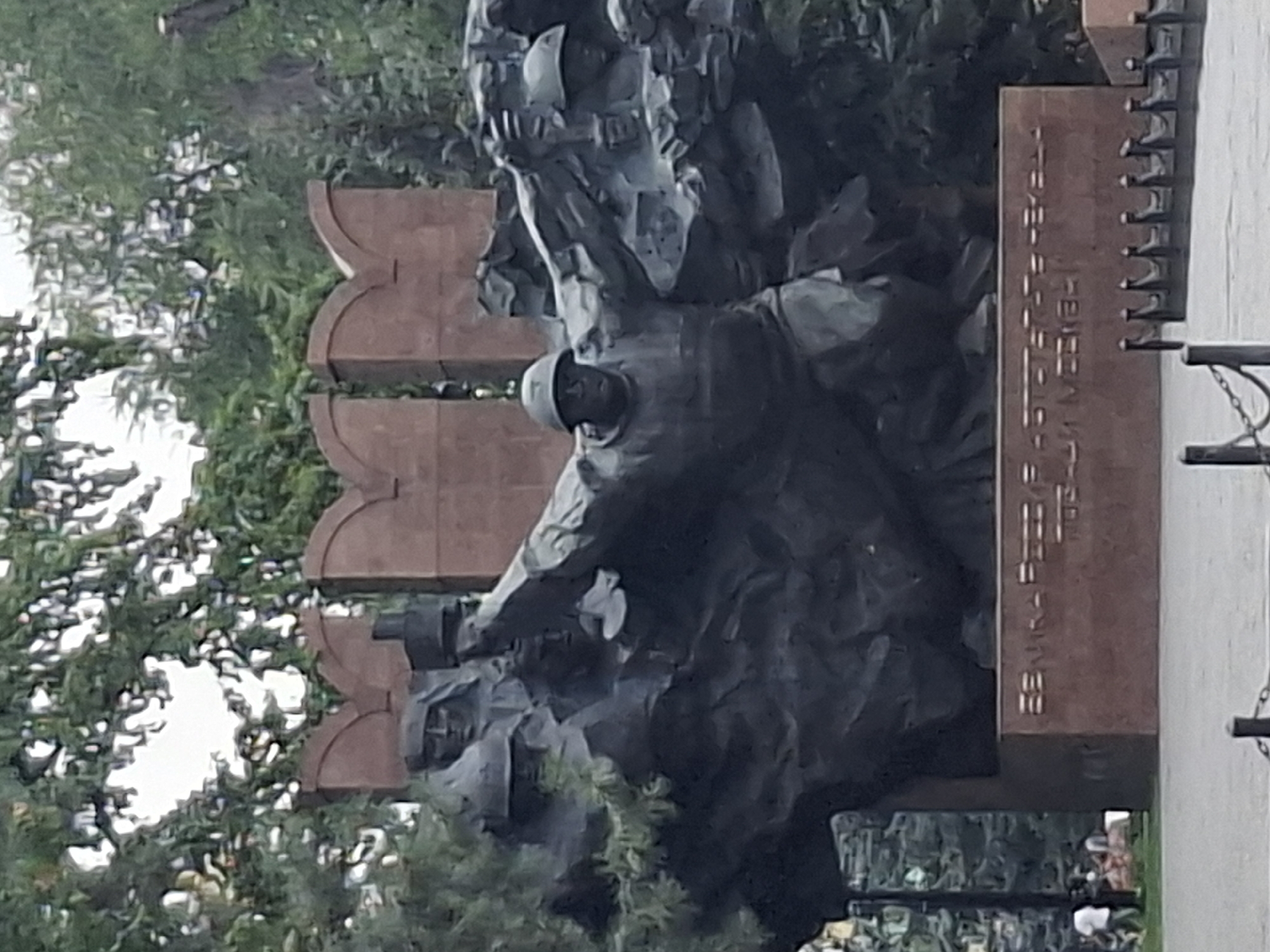 The image shows the "Eternal Flame" monument in Almaty, Kazakhstan, dedicated to soldiers who died in World War II. The monument features a large, dark bronze sculpture of soldiers in combat, with one soldier prominently in the foreground, arm raised as if throwing a grenade. Behind him, other figures emerge from stylized rock formations. The sculpture is mounted on a reddish-brown pedestal with Cyrillic text inscribed on its front face. In the background, lush green trees create a natural setting. To the left of the monument, a few people, including a person in a white t-shirt, are visible walking in a park-like area. A chain-link fence lines the front of the monument, with a few posts visible. The overall scene depicts a solemn memorial site.