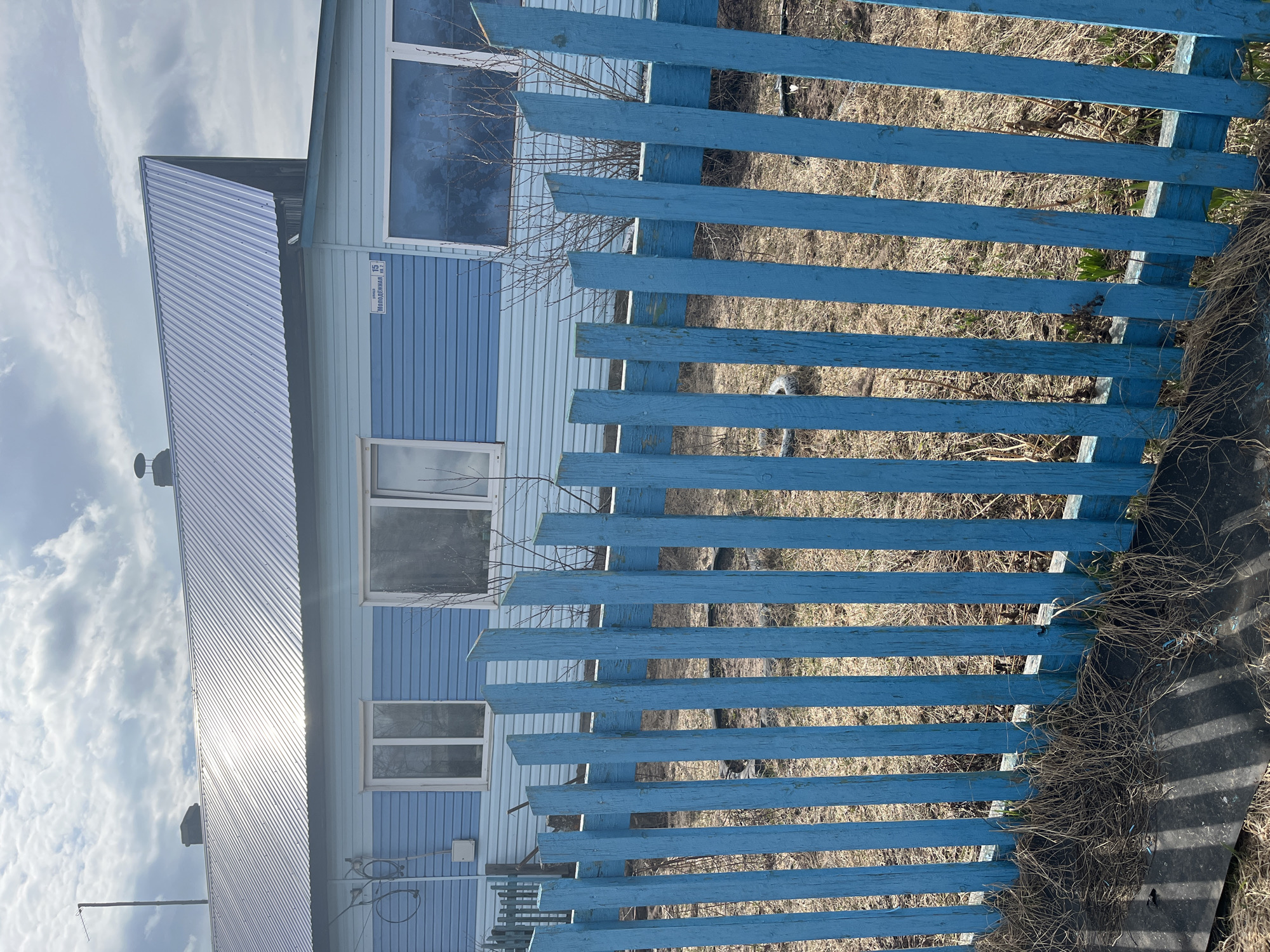 The image displays a static scene featuring a light blue building and a weathered blue fence under a partly cloudy sky. The building, likely two stories, has horizontal siding in two shades of blue – a lighter tone on the lower section and a slightly darker one above. It features several rectangular windows with white frames. Its roof is made of corrugated metal, possibly silver or light gray, reflecting bright sunlight. A small white sign is visible on the building, bearing text that reads "улица Молодёжная, 15" (Molodyozhnaya Street, 15).
In front of the building, a wooden picket fence, painted a vibrant but somewhat faded blue, stands prominently. The fence consists of horizontal planks, through which dry, sparse grass and bare tree branches are visible, suggesting a late winter or early spring setting. To the far right, near the ground level, there's an area with dried grass and some dark material, possibly a tarp, casting shadows. The overall impression is of a quiet daytime scene in a rural or suburban environment, consistent with the provided location context of Буренка, Russia.