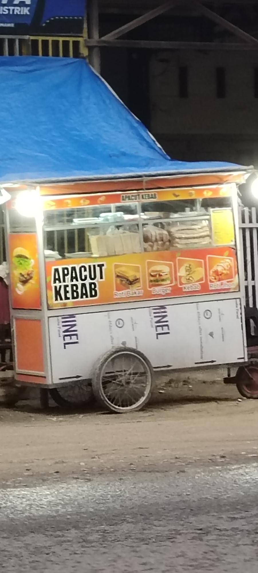 At night in Lhokseumawe, Indonesia, a brightly lit "APACUT KEBAB" food cart stands outdoors, likely operating by a roadside. The cart is covered by a blue tarp, providing shelter. The front of the cart features large signage with "APACUT KEBAB" in prominent letters and images of the food offered: "Roti Bakar" (grilled bread), "Burger", "Kebab", and "Roti Jhon". Inside the glass display, various food ingredients like bread and patties are visible. The lower section of the cart is made of white panels, with the word "MINEL" and what appears to be "Alumunium Panel" printed on them, along with a recycling symbol. The cart has a large spoked wheel on the left and a smaller, red-colored wheel on the right, suggesting it is mobile. The ground is unpaved, appearing to be dirt or gravel. In the dark background, structural elements like wooden beams and some yellow railings are visible, along with a partially obscured sign that reads "ISTRIK" (likely part of "LISTRIK" meaning electricity). The scene captures a typical street food vendor setting after dark.
