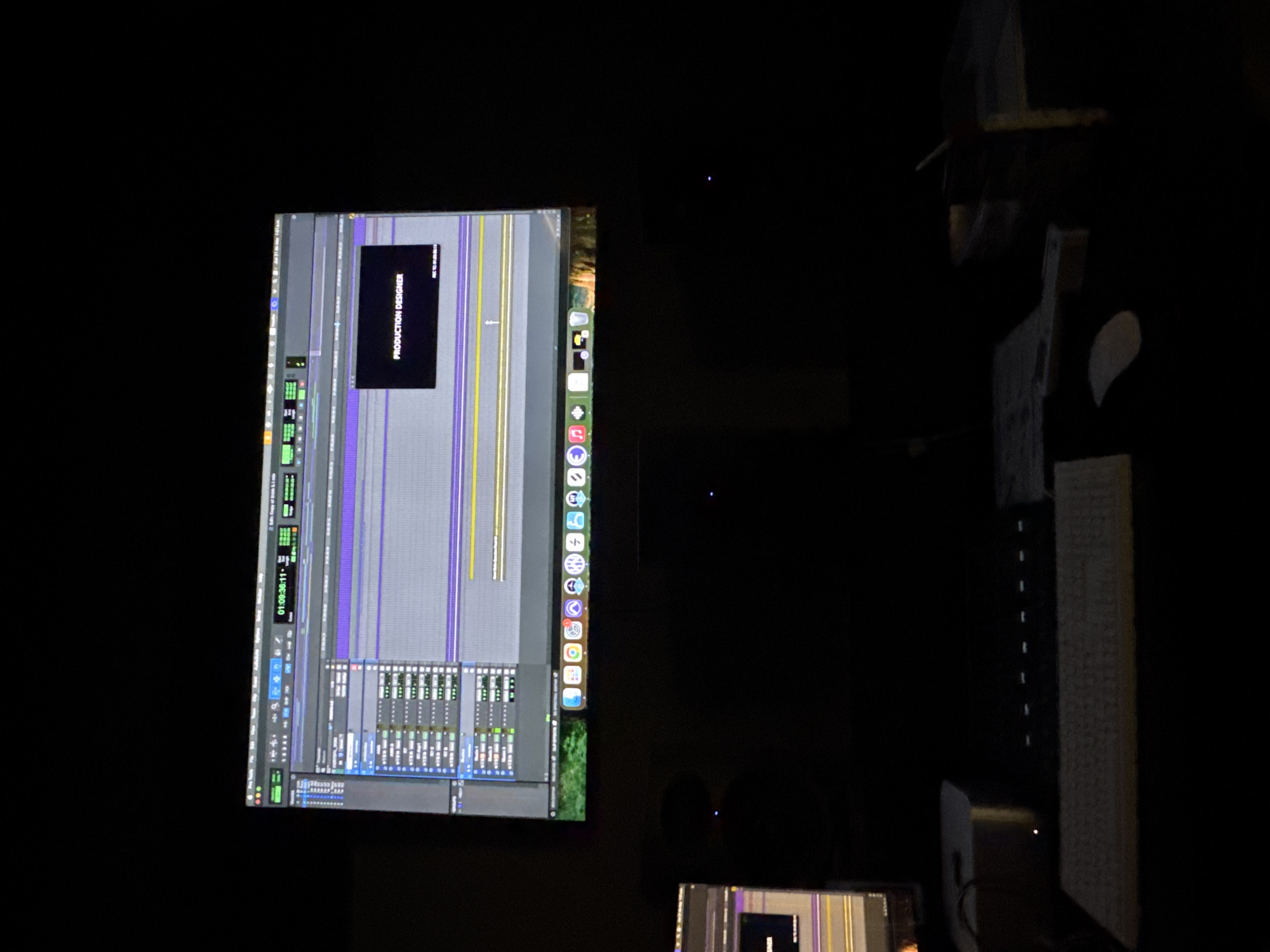 The image depicts a dimly lit workspace. A computer monitor displays a timeline-based audio editing interface, possibly a DAW like Pro Tools or Logic Pro. The interface shows multiple audio tracks with waveforms, faders, and other editing tools. A black box with the words "PRODUCTION DESIGNER" written in it are displayed on the upper side of the screen. Next to the monitor, on the right side of the image, there's a desk with a keyboard, a mouse, a desk lamp and other assorted items. Another monitor with the timeline editing interface is visible at the bottom of the scene. The location is unknown and there is no way to know if this photo was taken in Mexico City, Mexico or anywhere.