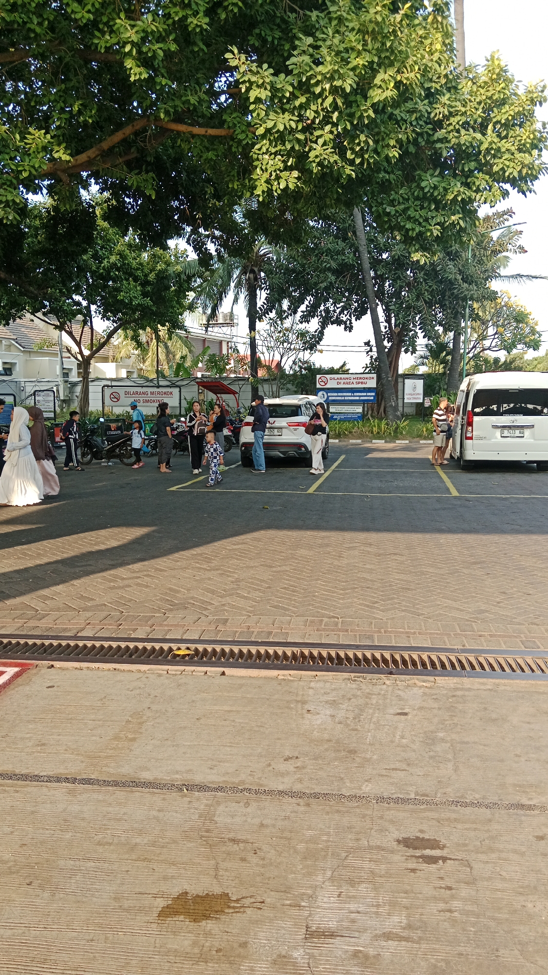 The image depicts a parking lot area in Banyuglugur, Indonesia, under the shade of large trees with lush green foliage. The ground is paved with a cobblestone-like pattern in the foreground, transitioning to a lighter colored concrete in the background where vehicles are parked. A metal grate runs along the edge of the cobblestone area.

Several people are present in the parking lot. On the left, two individuals wearing white and brown hijabs are walking. To their right, a group of people, including children, are gathered near motorcycles. One child, dressed in a checkered outfit, is walking alone in the middle of the parking lot.

There are multiple vehicles visible. A white SUV is parked in the center, and a white van is parked on the right side of the image. The white SUV has a license plate reading "3265 GS". The white van has a license plate reading "D 7433 AU". Two motorcycles are parked near a group of people.

Signs are also visible in the background. One sign reads "DILARANG MEROKOK NO SMOKING" in white text on a blue background, indicating a no-smoking area. Another sign, also blue and white, has text that appears to be Indonesian and English, likely providing additional regulations.

The environment suggests an outdoor public space, possibly a commercial area or a rest stop, given the presence of vehicles and signage. The lighting indicates it is daytime, with shadows cast by the trees and vehicles.