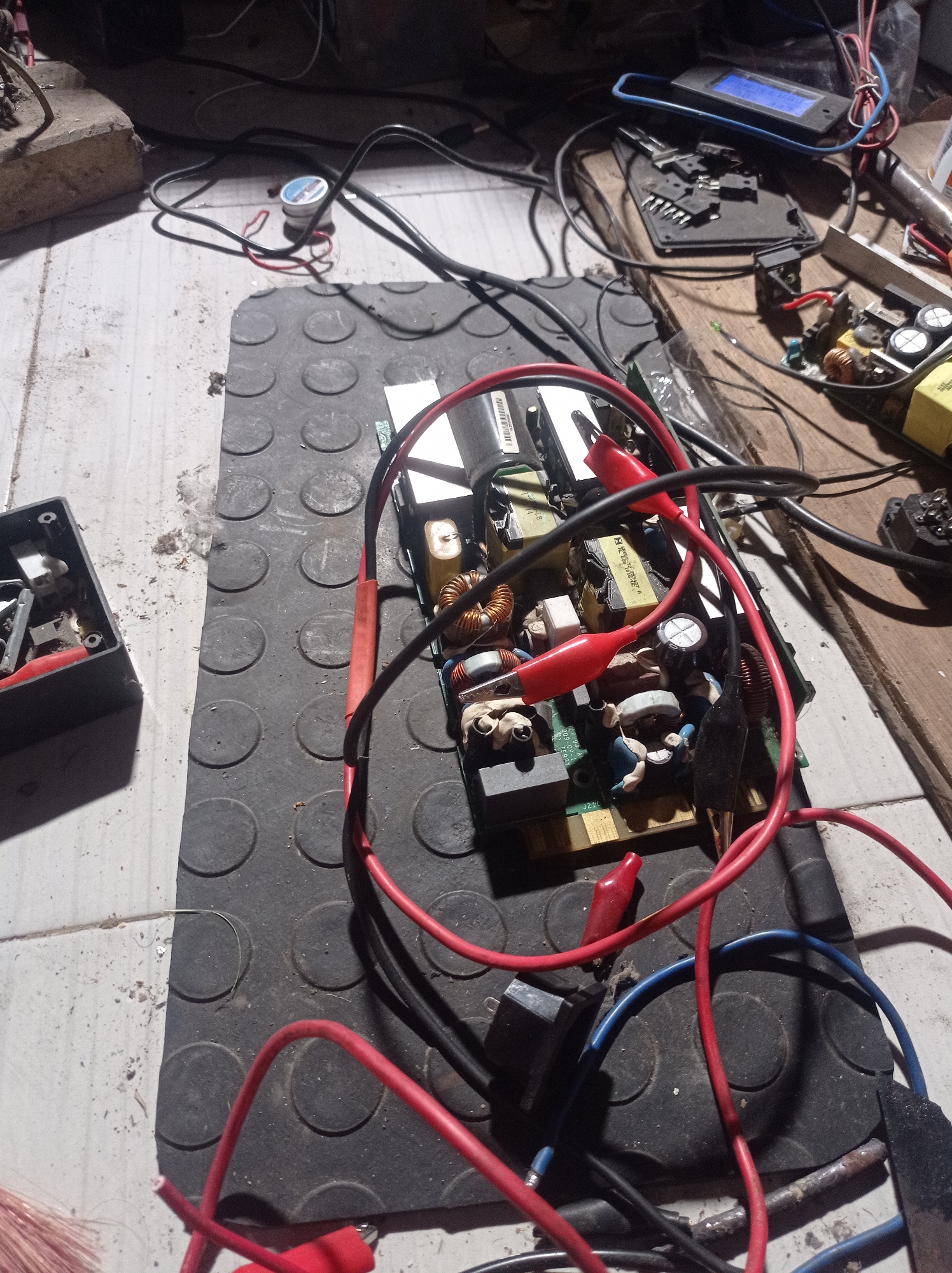 The image shows a cluttered workbench with electronic components and wires. A circuit board, possibly a power supply, is the central focus, connected to various wires with alligator clips. Various tools and electronic parts are scattered around, including what appears to be a digital multimeter. The environment is informal, with a textured mat and rough surfaces, suggesting a workshop or a DIY electronics setup. The presence of dust and debris indicates the location might be a workshop or garage. The scene does not provide any explicit clues about its location in Purwokerto, Indonesia.