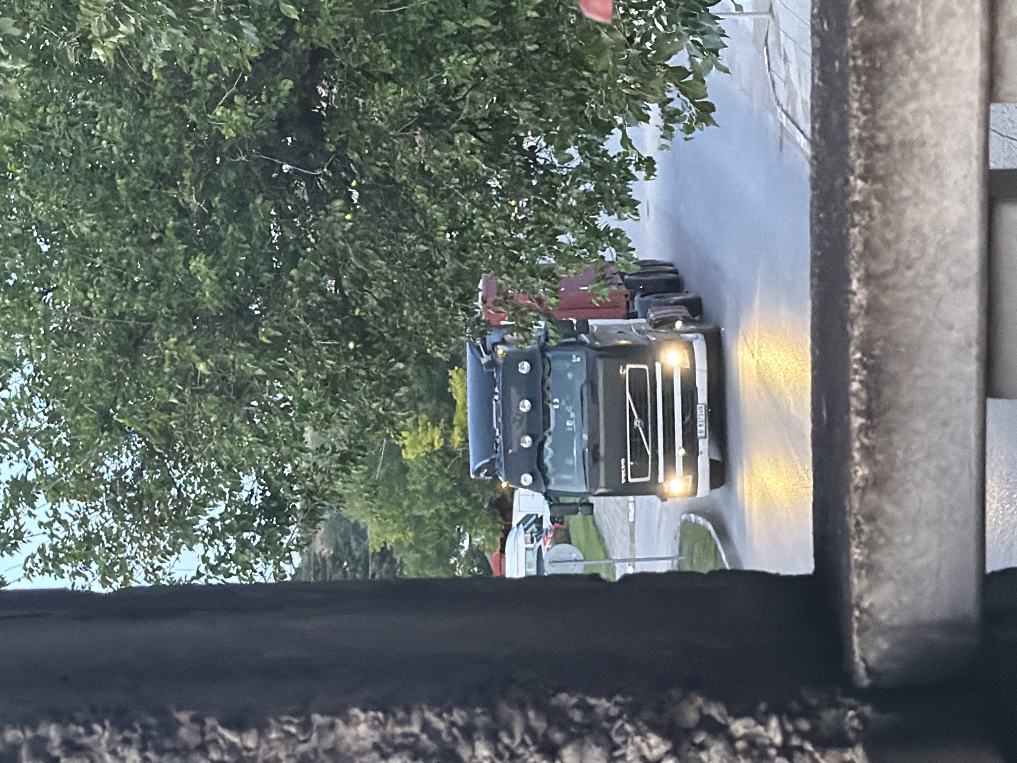 The image, oriented vertically, depicts a street scene with two large trucks at what appears to be dusk, night, or early morning, in Iskar, Bulgaria. The primary subject is a dark-colored (likely black or dark blue) Volvo semi-truck, its headlights brightly illuminated and facing to the right. The "VOLVO" brand name is distinctly visible on its front grille. Behind and slightly to the left of this truck, a second red truck is partially visible.
The trucks are situated on a paved surface that reflects the headlights, possibly indicating wet conditions or simply reflective asphalt. Abundant green foliage from trees fills the left side and background of the image, suggesting a tree-lined street or a wooded area adjacent to the road. A portion of a building or wall with a textured, stone-like surface frames the right side of the composition. No people are visible in the scene. Aside from the "VOLVO" branding, no other text is clearly legible. The overall atmosphere is quiet, with artificial light from the truck headlights piercing the low-light conditions.