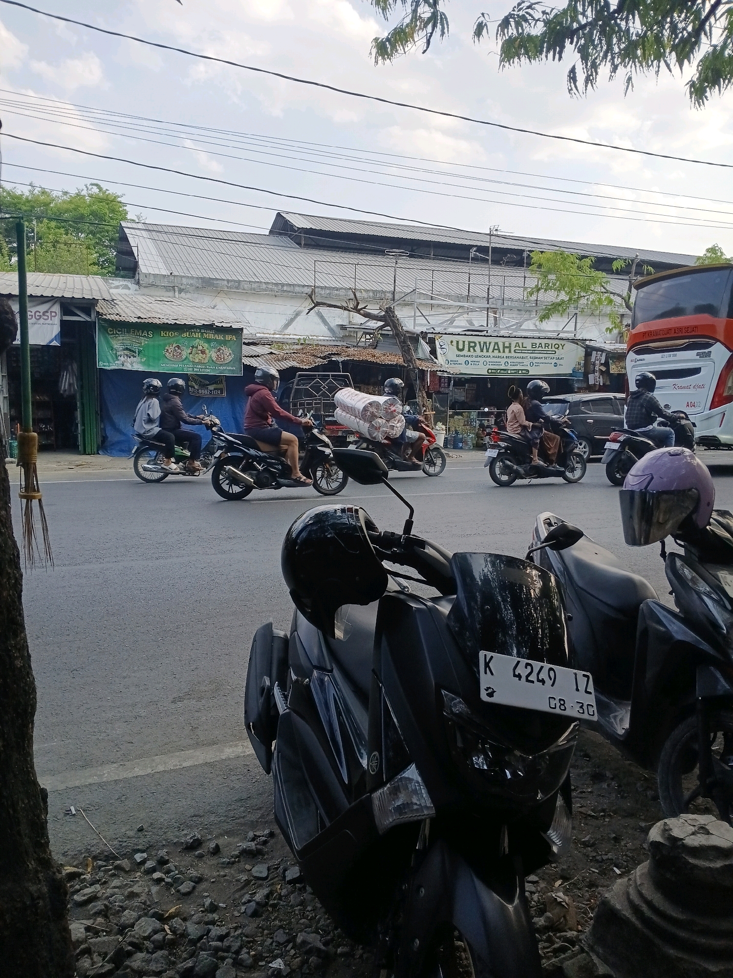 In Purwodadi, Indonesia, several motorcycles traverse a paved street. A motorcycle in the foreground is black, with a white license plate reading "K 4249 IZ 08.30". Behind it, another black motorcycle is visible. Further down the street, a black motorcycle carries two individuals wearing helmets, and another black motorcycle transports a single rider, also helmeted, with a large roll of white material strapped to its back. To the right, a red and white bus is parked. On the left side of the street, there are buildings with awnings and signs, one reading "URWAH AL BARIQY". The sky is cloudy, and trees are visible in the background. The area is lined with rocks and dirt on the side of the road.