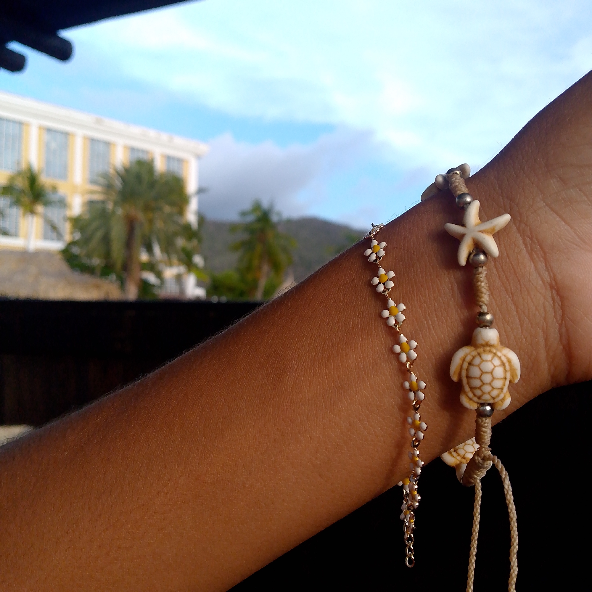 The image provides a close-up view of a person's arm and wrist, adorned with two distinct bracelets, set against a blurred tropical background. The skin on the arm appears to be a warm brown tone.
One bracelet is delicate, featuring a chain with small white beads that have yellow centers, resembling daisies. The second bracelet is more substantial, made from a light beige woven cord. It incorporates silver-colored beads and two prominent charms: a white starfish and a white sea turtle. Both bracelets are themed with elements commonly associated with beaches and nature.
In the softly focused background, a large, multi-story building, likely a resort or hotel, can be seen. It features light yellow walls and numerous windows with white frames. Several palm trees are visible around and behind the building, reinforcing the tropical setting. Distant mountains or hills are silhouetted against a bright, cloudy blue sky. A dark, out-of-focus element in the foreground suggests a railing or table from which the photo was taken.
The scene conveys a relaxed, daytime atmosphere, consistent with a vacation or leisure setting in a tropical region like Pedro González, Venezuela. There is no visible text in the image.